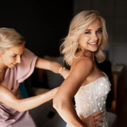 Panna młoda w sukni ślubnej z koronkowymi aplikacjami, poprawiana przez druhnę w pudrowej sukience, uśmiechając się do kamery.