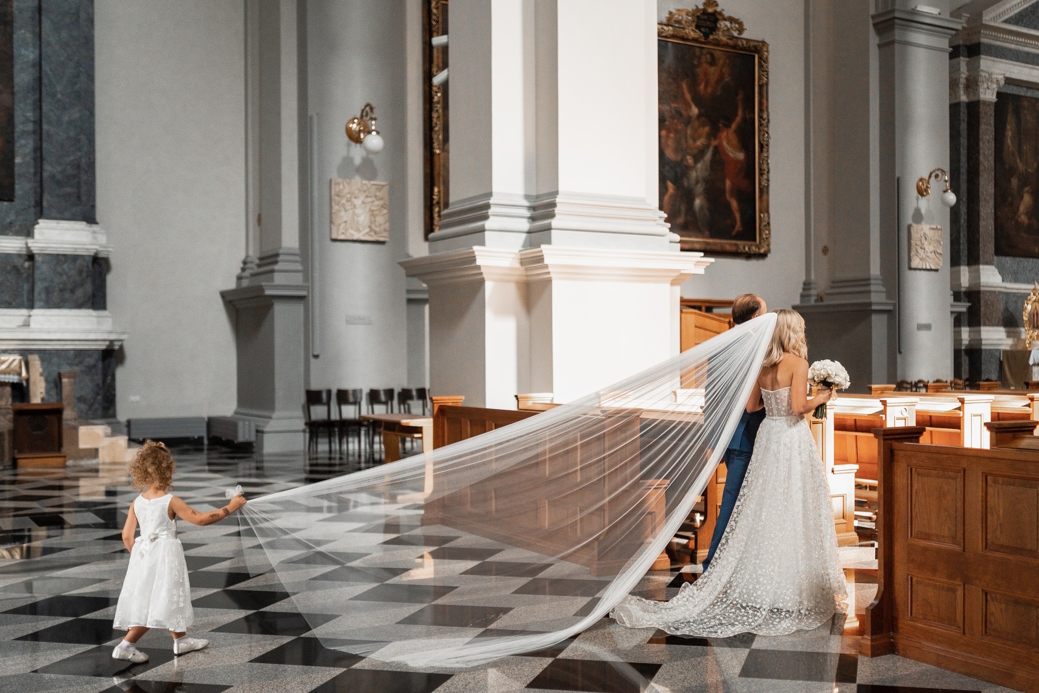Panna młoda z bukietem i pan młody w kościele, dziewczynka trzyma długi welon na czarno-białej podłodze, w tle kolumny i obrazy.