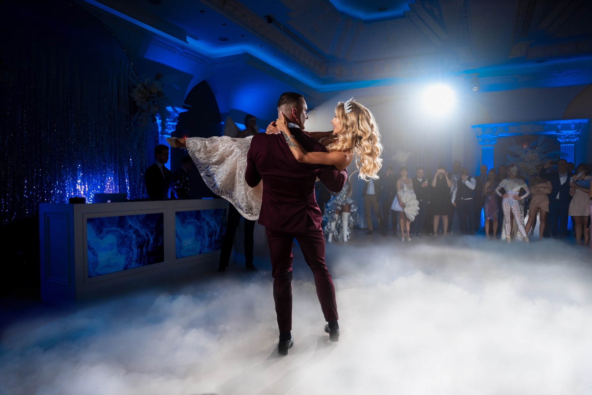 Para młoda w trakcie pierwszego tańca w Warszawie, panna młoda w koronie trzymana w ramionach przez pana młodego w burgundowym garniturze, tło wypełnione gośćmi i dekoracjami podświetlonymi...