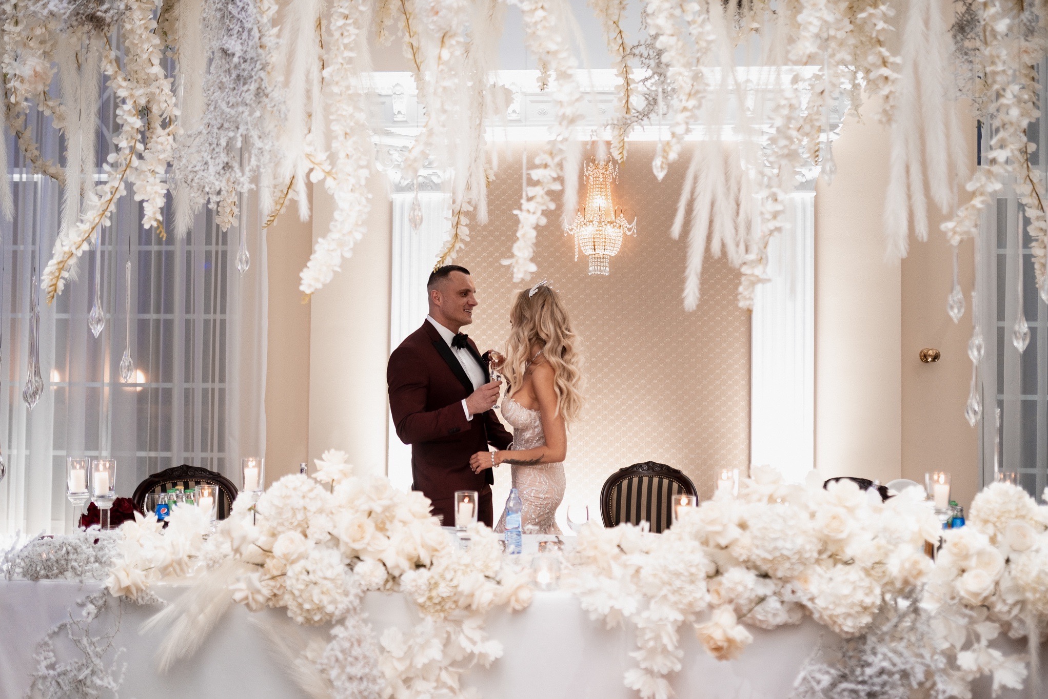 Para młoda w eleganckich strojach wznosi toast przy bogato zdobionym stole weselnym z kwiatami i zwisającymi dekoracjami, oświetlona kryształowym żyrandolem.