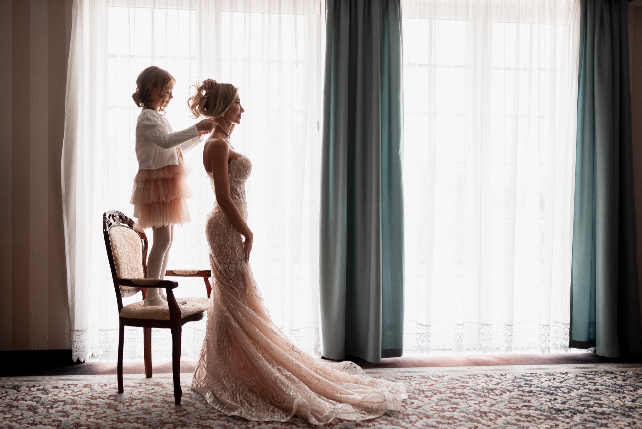 Panna młoda w długiej, zdobionej sukni, profil, dziewczynka stojąca na krześle zapina jej naszyjnik, jasne okno w tle.