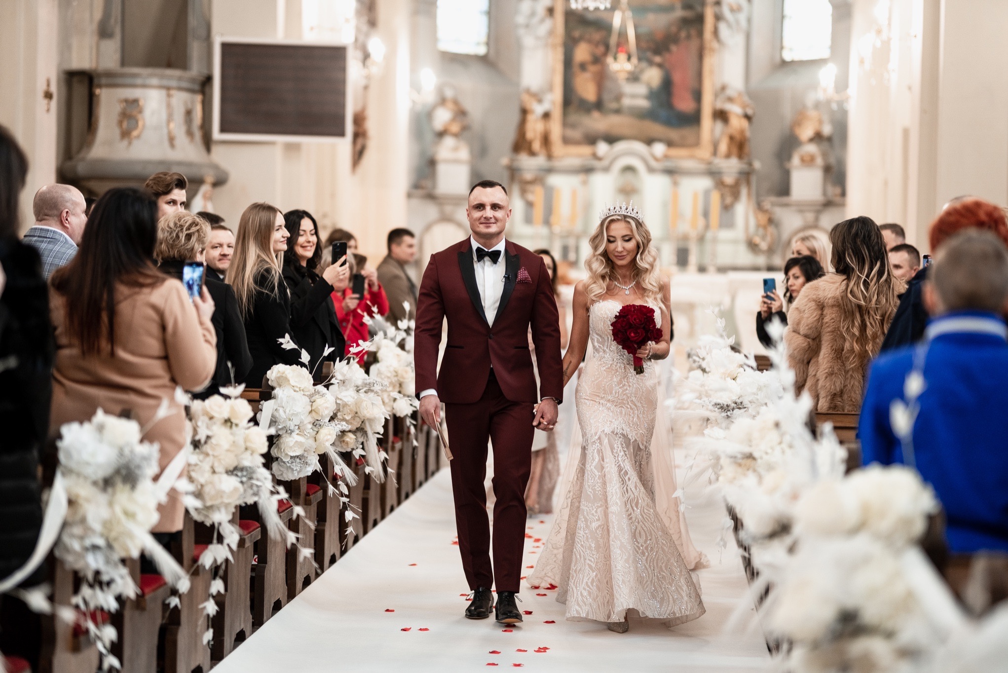 Para młoda w bordo garniturze i koronkowej sukni przechodzi alejką udekorowaną białymi kwiatami w kościele, goście robią zdjęcia telefonami.