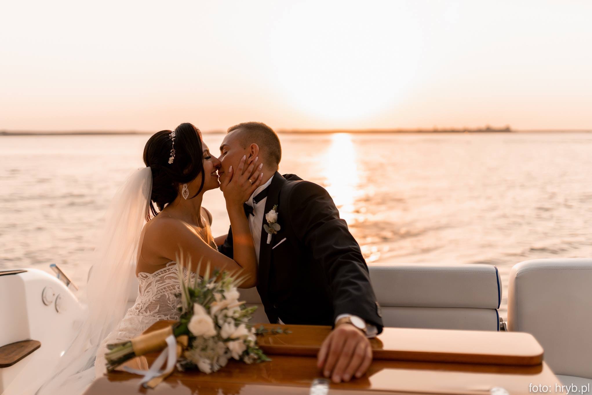 Pocałunek nowożeńców na łodzi podczas zachodu słońca, bukiet ślubny na drewnianym blacie, ona w koronkowej sukni, on w smokingu z białą butonierką.