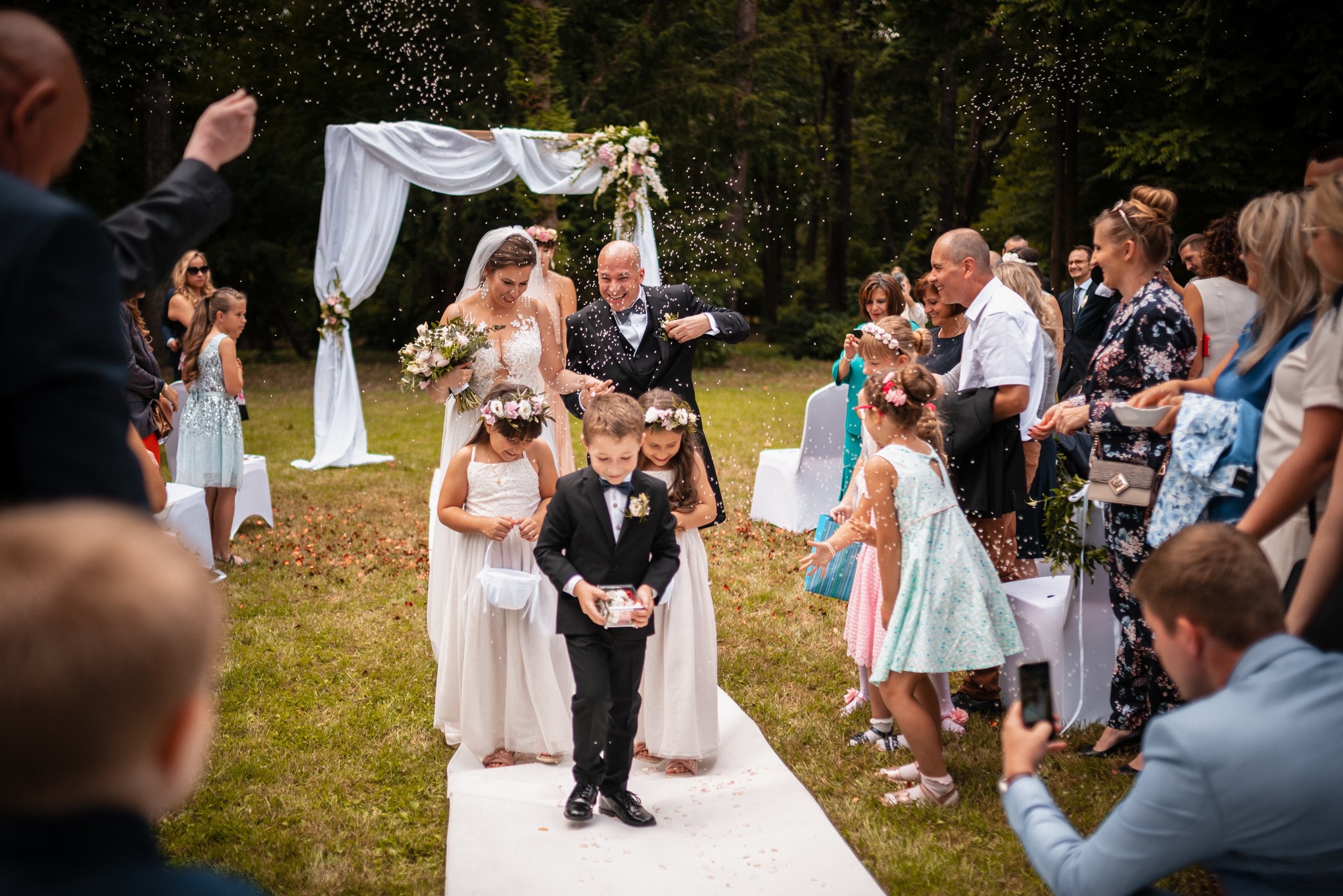 Para młoda przechodzi pod białym łukiem udekorowanym kwiatami, obsypywana konfetti przez gości i dzieci niosące poduszki z obrączkami na ślubie plenerowym.