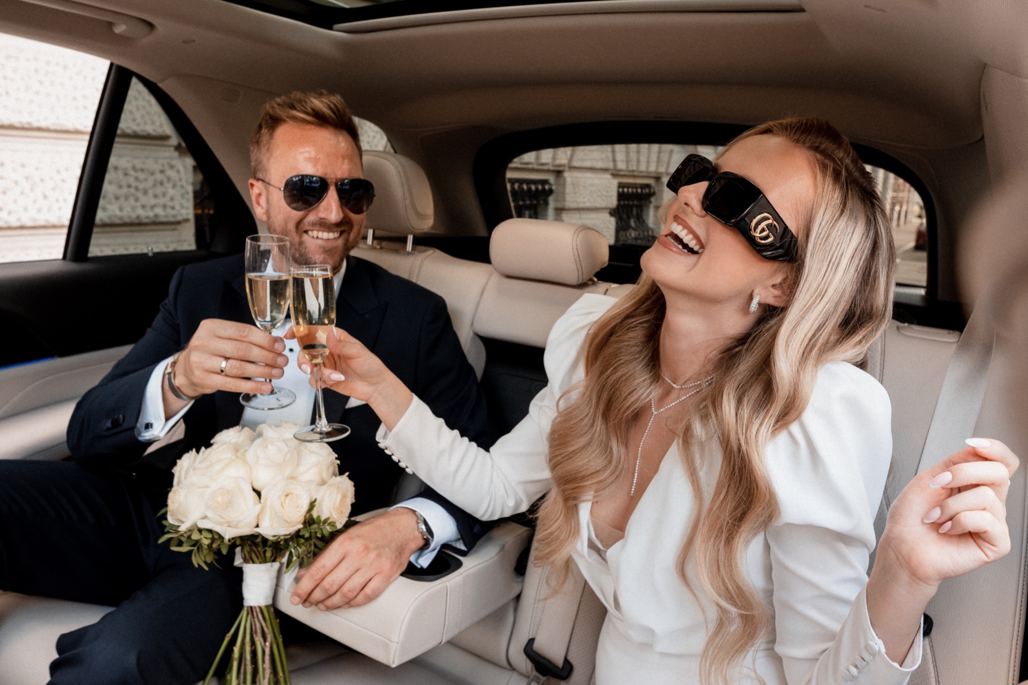 Uśmiechnięta para młoda w samochodzie wznosi toast szampanem, panna młoda w dużych okularach przeciwsłonecznych Gucci, bukiet białych róż na kolanach pana młodego.