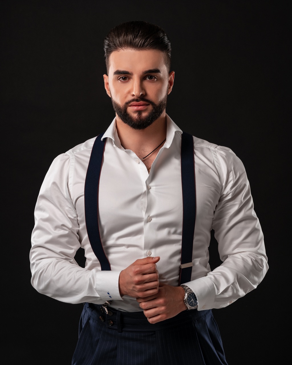 Portret mężczyzny z brodą w białej koszuli i szelkach na ciemnym tle, z zegarkiem na nadgarstku i splecionymi palcami.