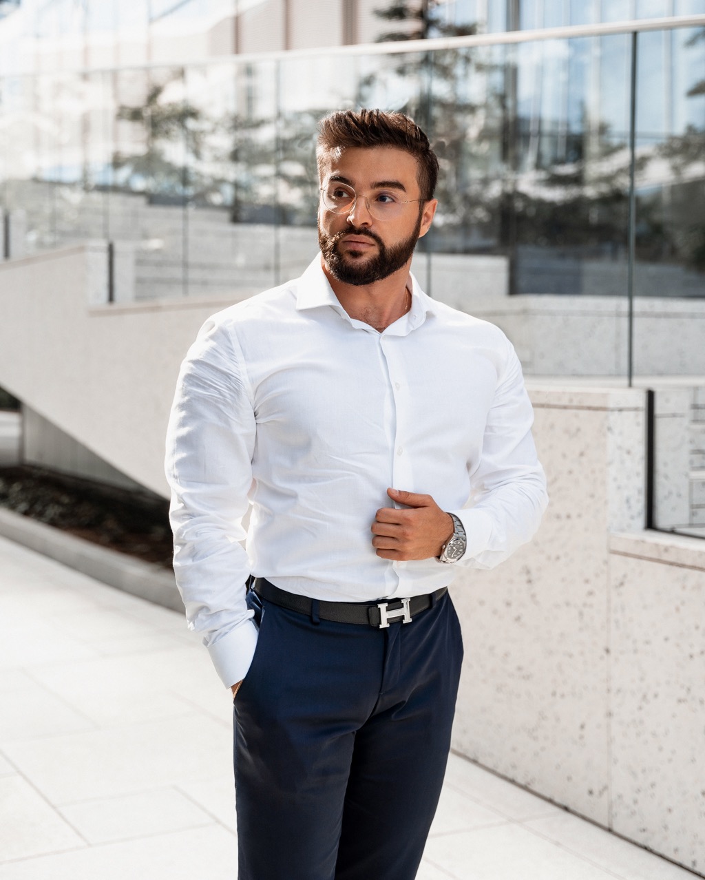 Mężczyzna w białej koszuli i granatowych spodniach, z brodą i okularach, pozuje na tle nowoczesnego budynku z przeszkloną fasadą. Zegarek na nadgarstku i pasek z klamrą w kształcie litery H.