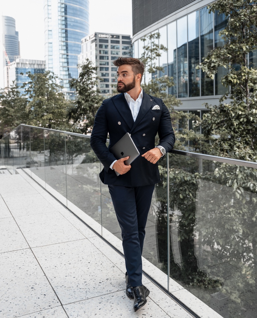 Mężczyzna w granatowym garniturze, białej koszuli i okularach, trzymający laptopa, na tle nowoczesnej architektury miejskiej.