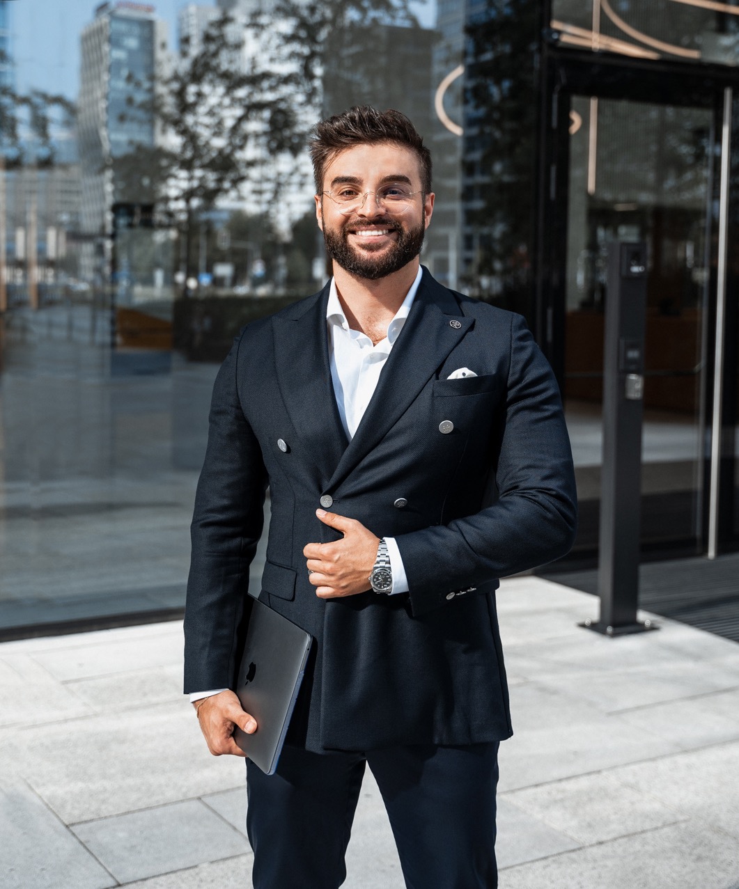 Uśmiechnięty mężczyzna w dwurzędowym granatowym garniturze, białej koszuli i okularach, trzymający laptopa na tle nowoczesnego budynku biurowego.