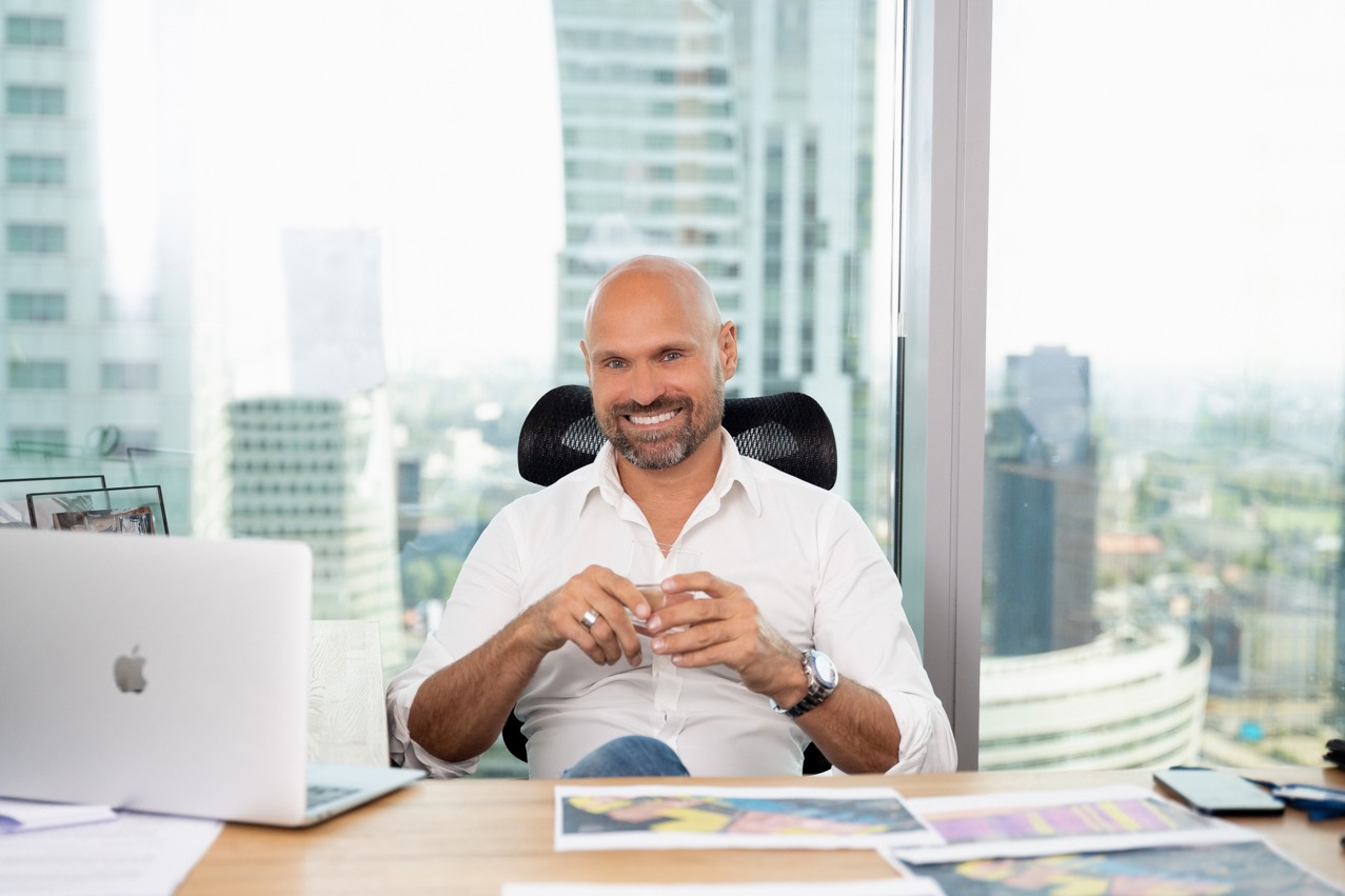 Uśmiechnięty mężczyzna w białej koszuli siedzi za biurkiem w nowoczesnym biurze z widokiem na miasto, z laptopem Apple i rozłożonymi szkicami na blacie.