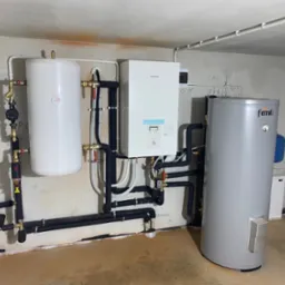 Zestaw urządzeń grzewczych: biały zbiornik, jednostka Panasonic Aquarea i szary zasobnik Ferroli, połączone rurami w piwnicy z surowymi ścianami.