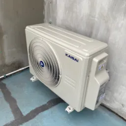 Zewnętrzna jednostka klimatyzacji Kaisai zamontowana na betonowej posadzce z widocznymi śladami po malowaniu, przy szarej, surowej ścianie.