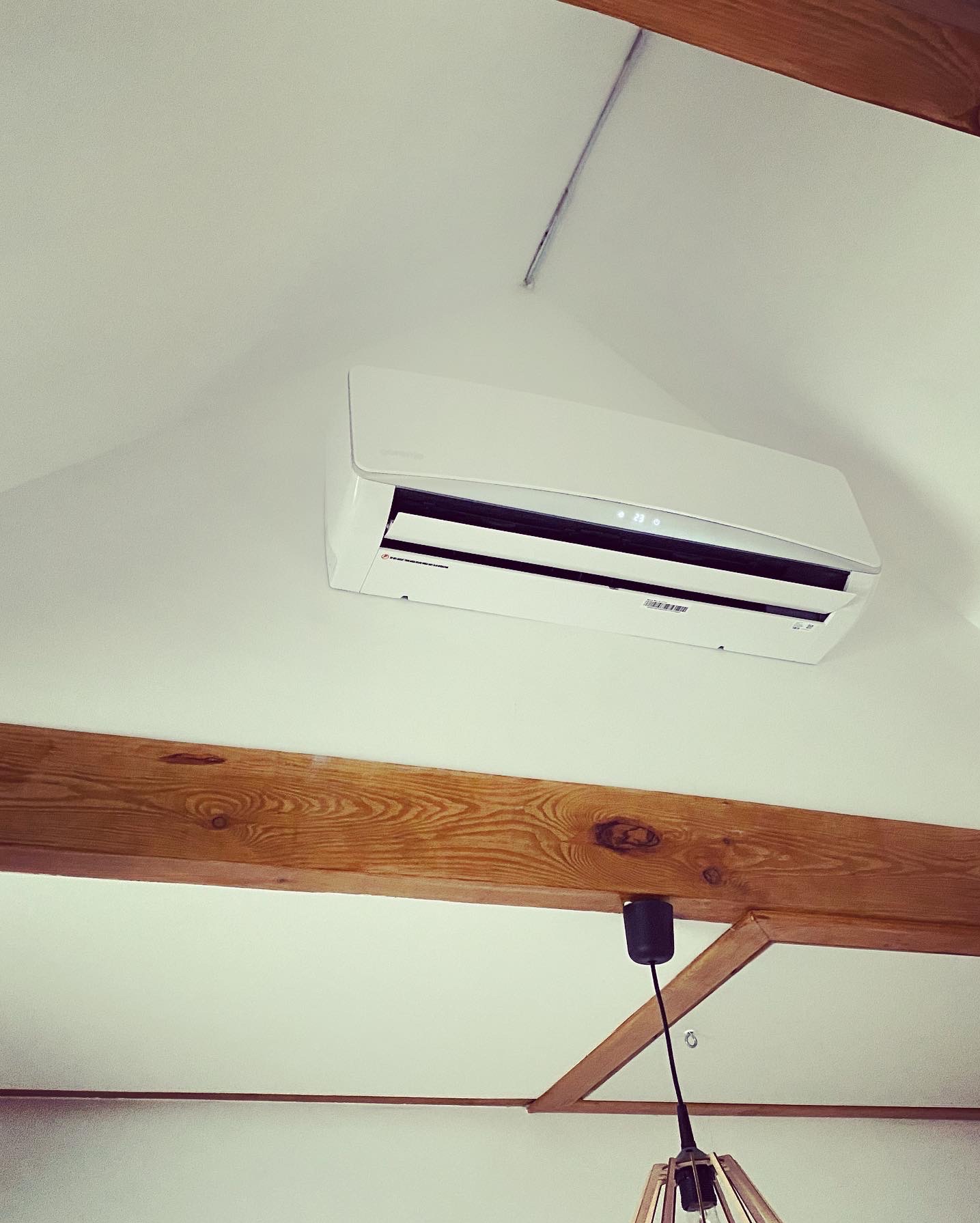 Biały klimatyzator zamontowany na suficie w pomieszczeniu z drewnianymi belkami stropowymi i częściowo widoczną lampą wiszącą.