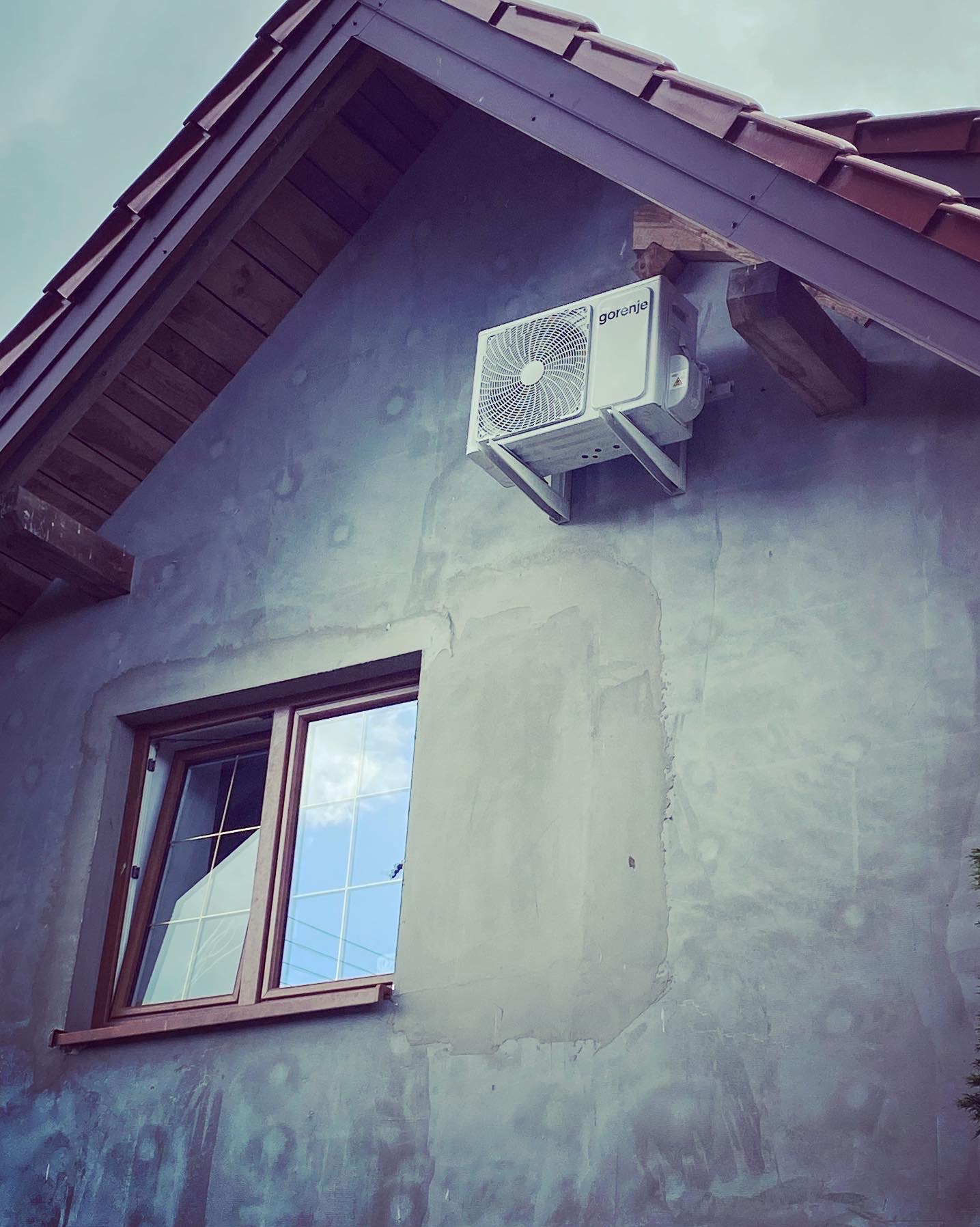 Biała jednostka klimatyzacyjna marki Gorenje zamontowana na szarej, surowej elewacji domu z drewnianymi elementami pod dachem i oknem z odbiciem nieba.