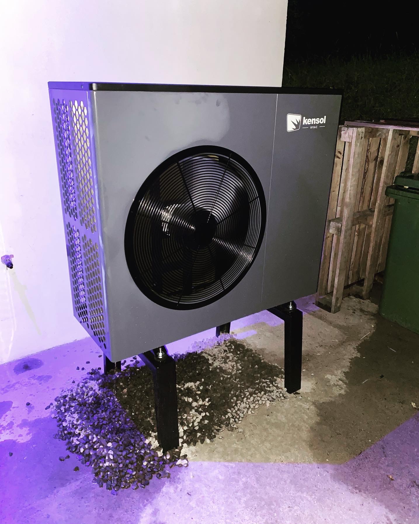 Szara pompa ciepła Kensol na czarnych nogach, ustawiona na żwirze obok ściany i palety, oświetlona fioletowym światłem.