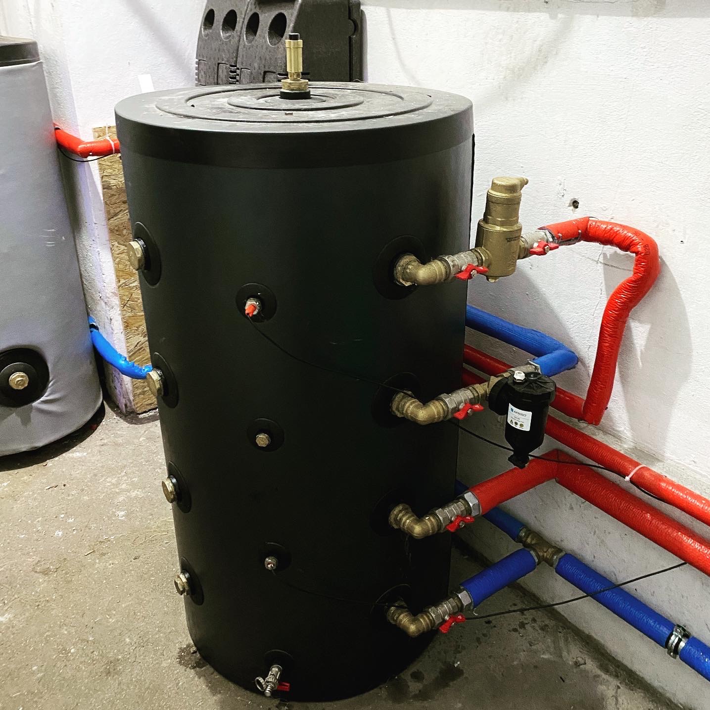 Czarny zbiornik akumulacyjny z licznymi przyłączami rur w kolorach czerwonym i niebieskim, zamontowany w kotłowni z widocznym filtrem na rurze i drugim zbiornikiem w tle.