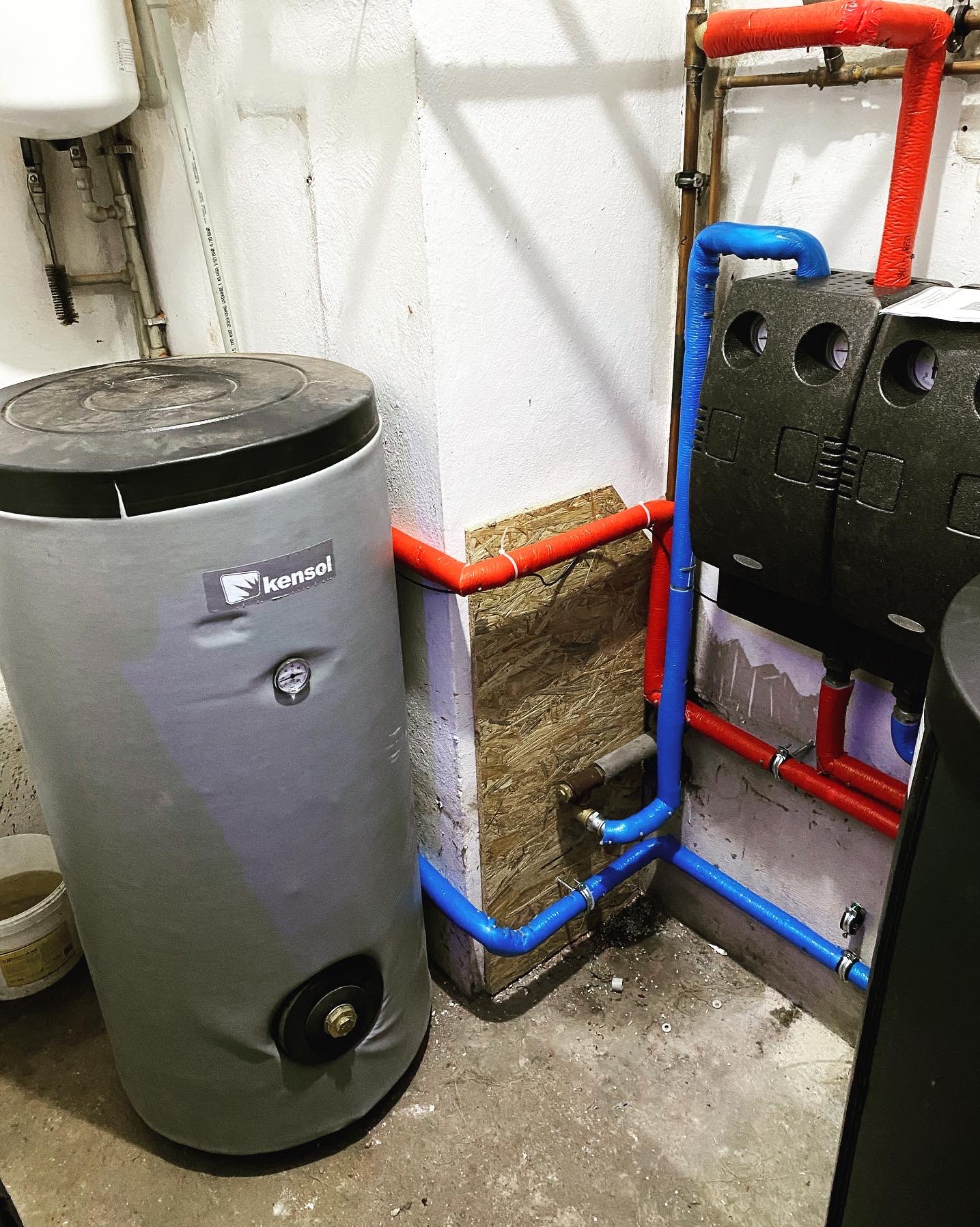 Szary zbiornik akumulacyjny Kensol z czarną pokrywą i manometrem, podłączony rurami w kolorze czerwonym i niebieskim, widoczne elementy instalacji grzewczej w piwnicy.
