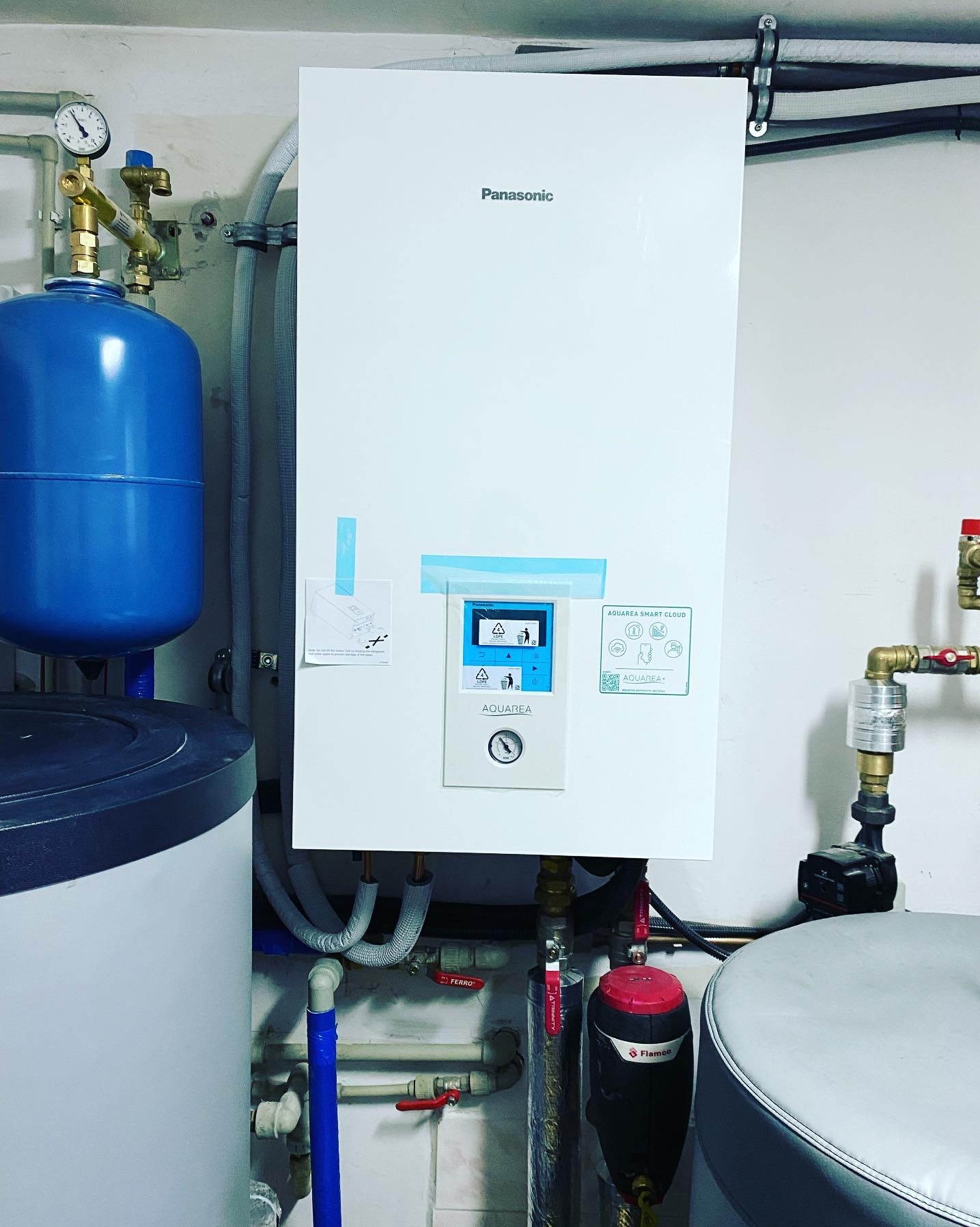 Biała pompa ciepła Panasonic Aquarea Smart Cloud zamontowana na ścianie w pomieszczeniu technicznym, widoczne zbiorniki buforowe, zawory, manometry i filtry.