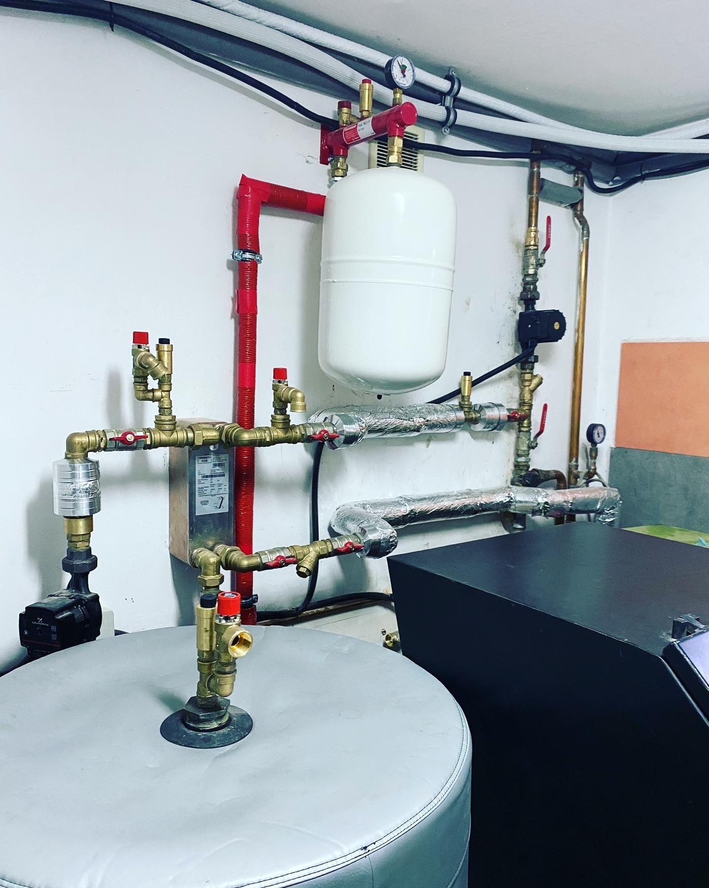 Złożona instalacja grzewcza z zbiornikiem akumulacyjnym, rurami miedzianymi, zaworami z czerwonymi pokrętłami i pompą obiegową w kotłowni, widoczne izolowane rury i manometr