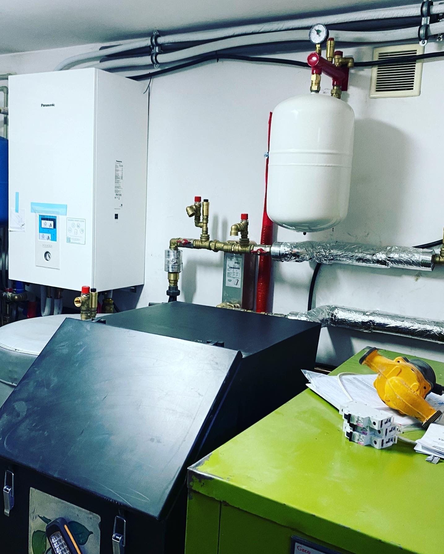 Biała pompa ciepła Panasonic z podłączonymi rurami i zbiornikiem wyrównawczym w pomieszczeniu technicznym, widoczne skrzynki narzędziowe i żółta pompa do odpowietrzania na zielonym blacie.
