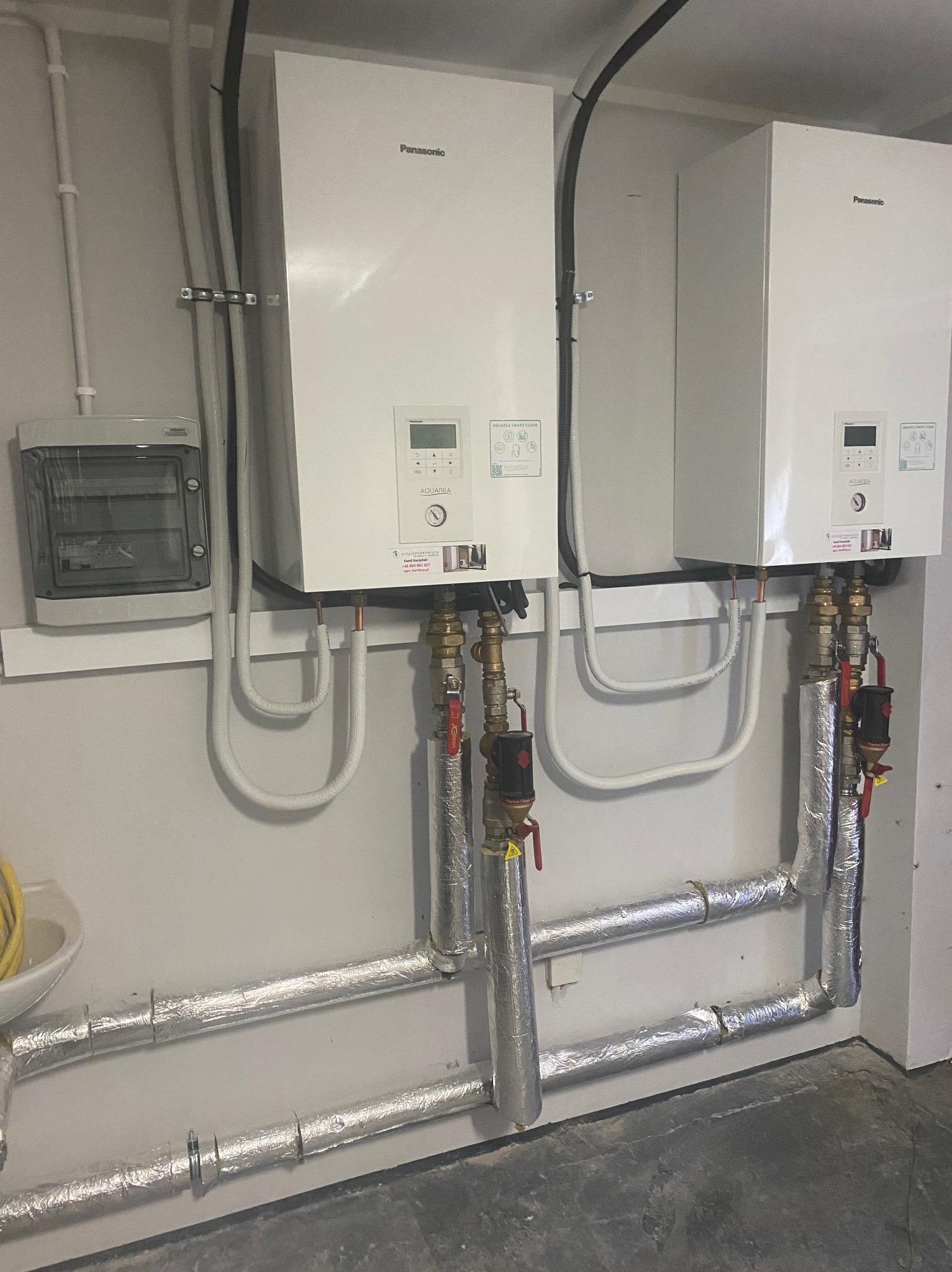 Dwa białe kotły grzewcze Panasonic Aquarea zamontowane na ścianie, z widocznymi rurami i zaworami, instalacja elektryczna i skrzynka bezpiecznikowa obok, rury izolowane folią aluminiową.