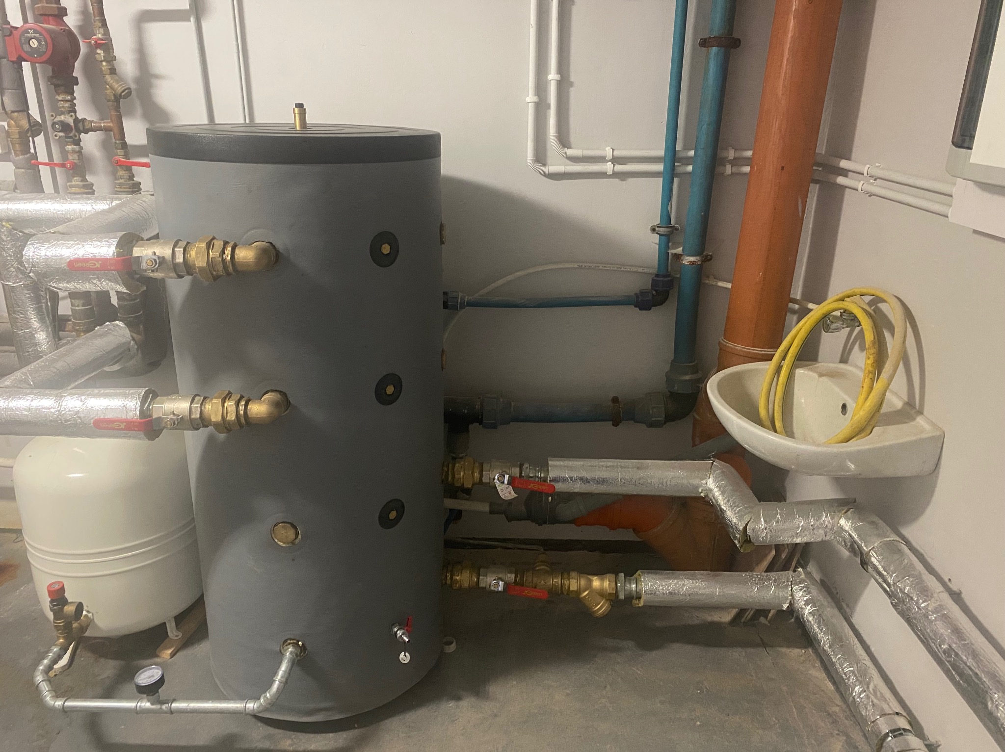 Szary zbiornik akumulacyjny ciepłej wody z przyłączonymi rurami miedzianymi i zaworami, biały zbiornik wyrównawczy obok, instalacja w pomieszczeniu technicznym z białymi ścianami i umywalką.