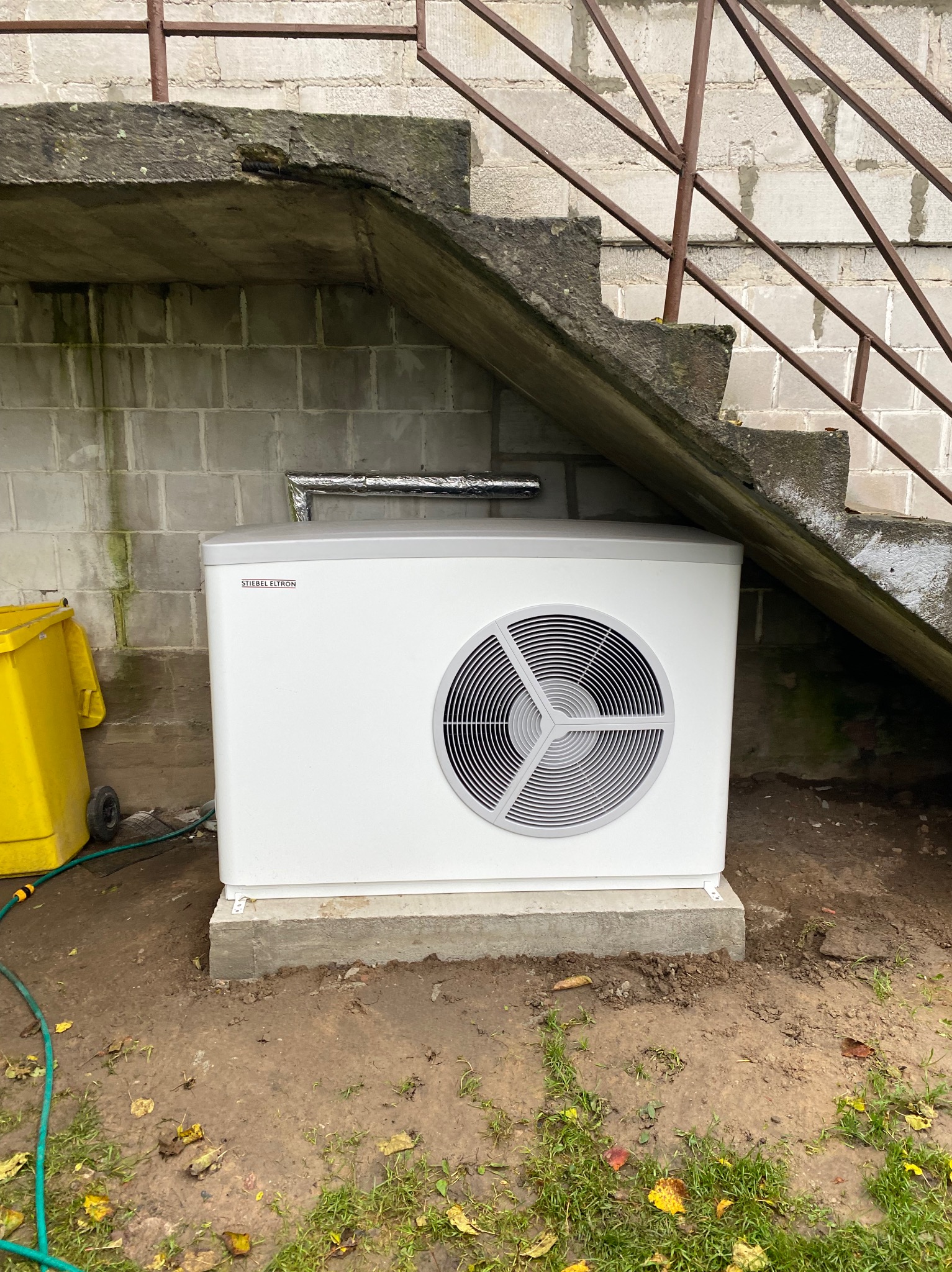 Biała pompa ciepła marki Stiebel Eltron ustawiona na betonowej podstawie przy ścianie budynku z betonowych pustaków i schodami z metalową barierką. Obok żółty kosz na śmieci i zielony wąż ogrodowy.
