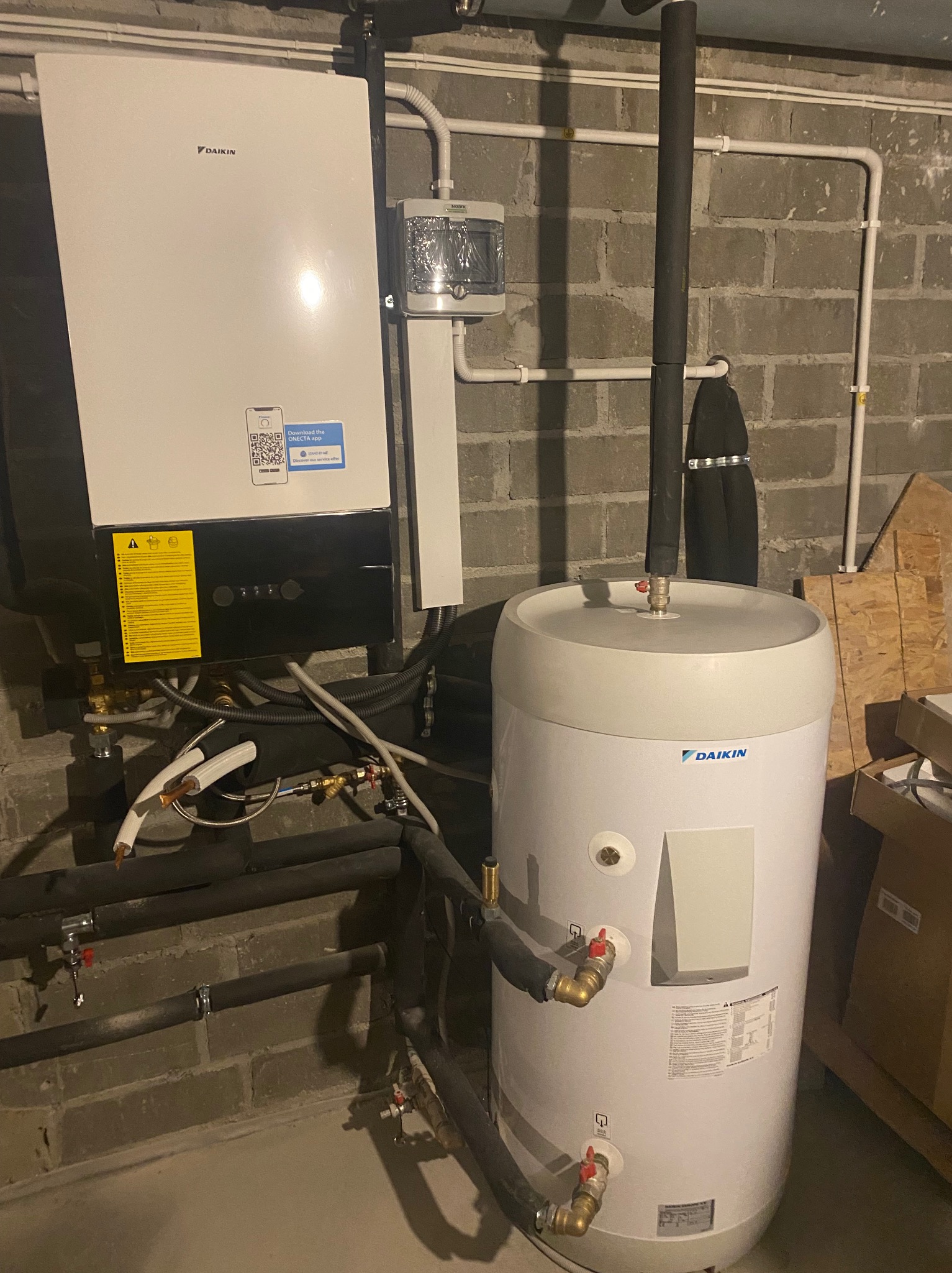 Biała pompa ciepła Daikin z podłączonymi rurami w kotłowni, widoczny zbiornik akumulacyjny, instalacja na tle ściany z cegły.