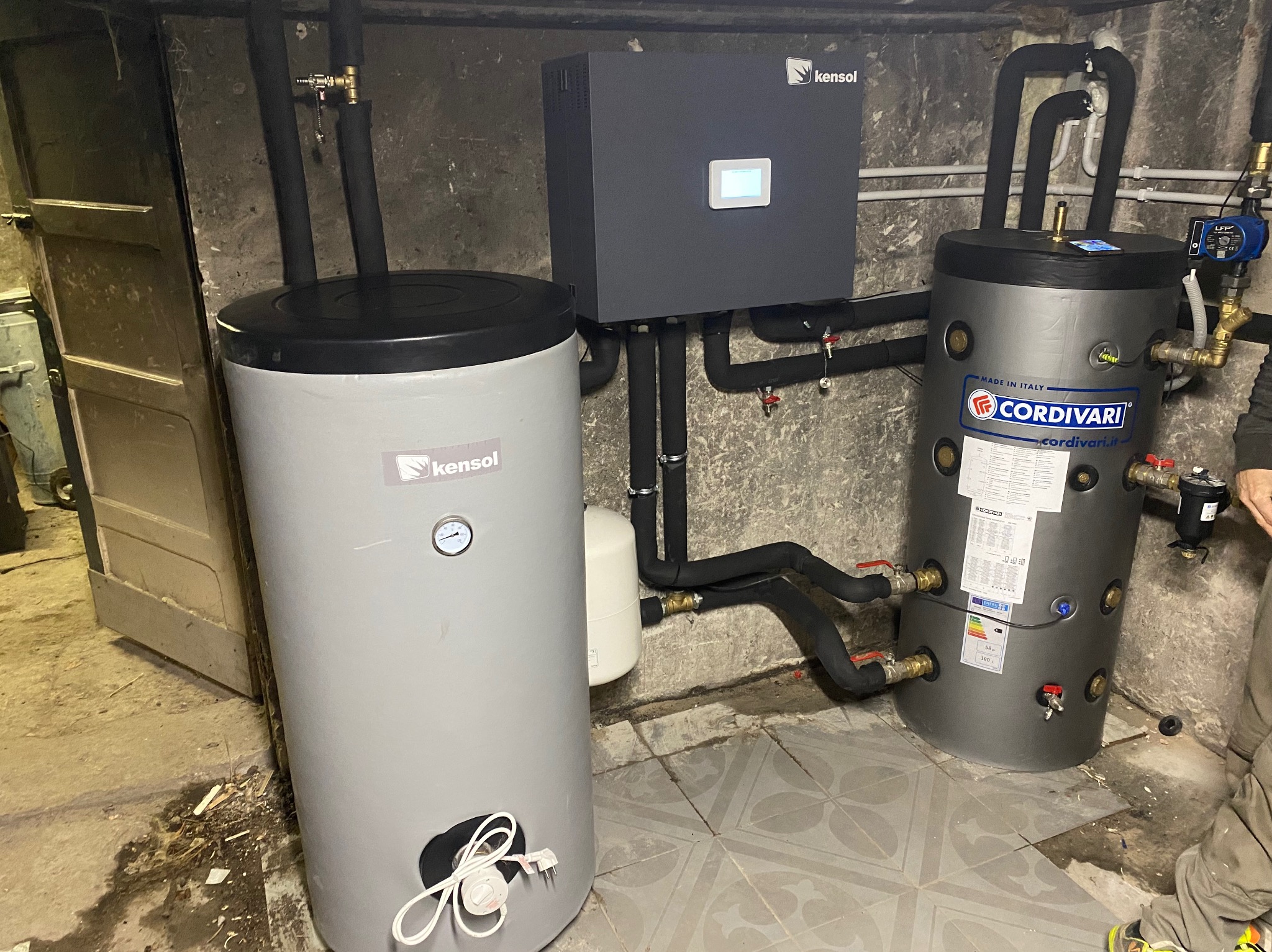 Dwa zasobniki ciepłej wody użytkowej marki Kensol i Cordivari połączone rurami, sterownik Kensol na ścianie, instalacja w piwnicy z betonowymi ścianami i płytkami na podłodze.