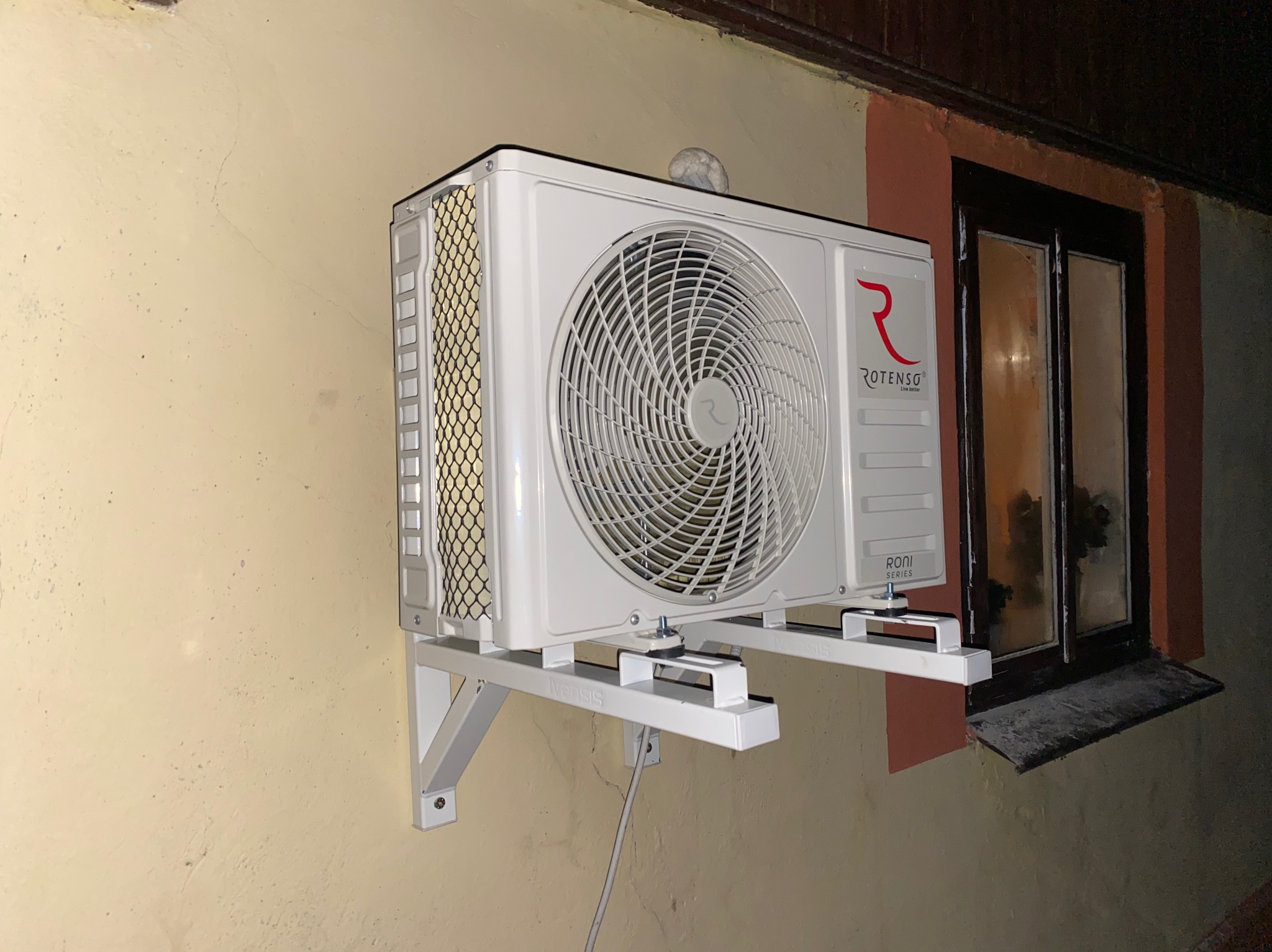 Biała jednostka zewnętrzna klimatyzatora Rotenso zamontowana na wspornikach na ścianie budynku obok okna z ciemną ramą. Na górze urządzenia leży niewielki kłębek kurzu.