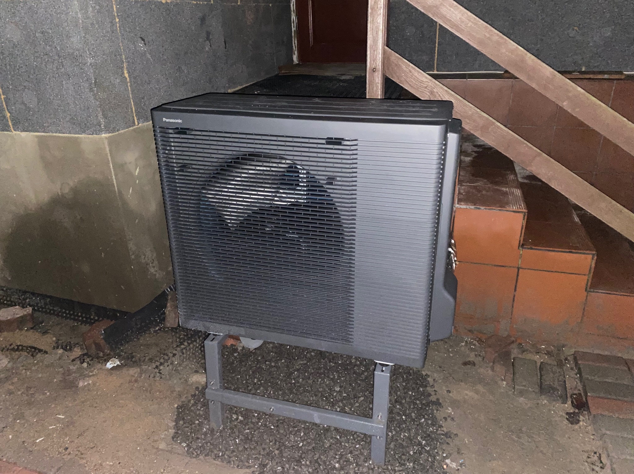 Zewnętrzna jednostka klimatyzacji Panasonic zamontowana na metalowej konstrukcji, widoczna na tle betonowej ściany i schodów.