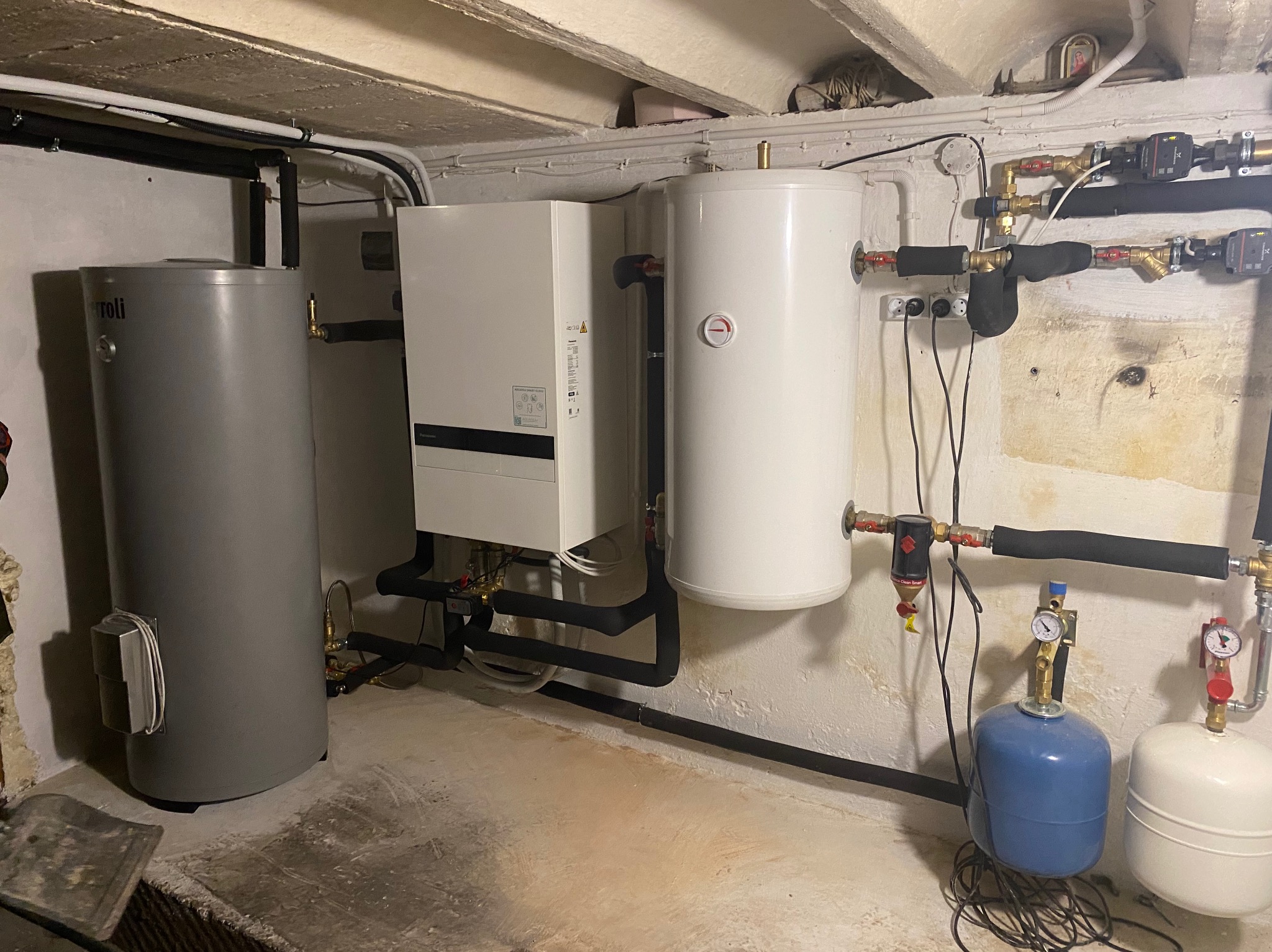 Zestaw urządzeń grzewczych w piwnicy: zbiornik akumulacyjny Ferroli, biały piec grzewczy, biały zasobnik ciepłej wody z termometrem, niebieski i biały zbiornik przeponowy oraz rury z zaworami.