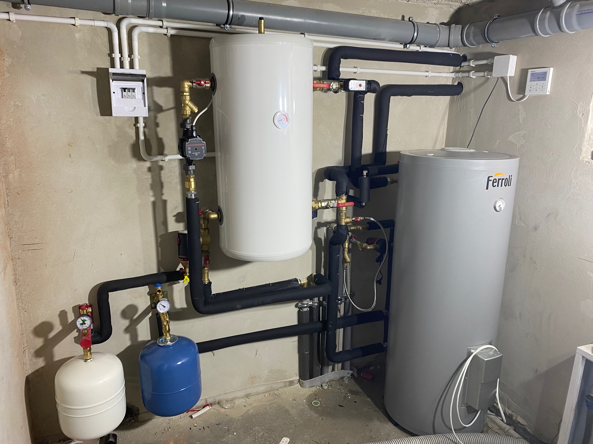 Kompleksowa instalacja grzewcza w kotłowni: dwa zbiorniki (biały i szary Ferroli), czarne rury z izolacją, manometry, zawory, rozdzielnica elektryczna na ścianie, widoczne elementy automatyki...