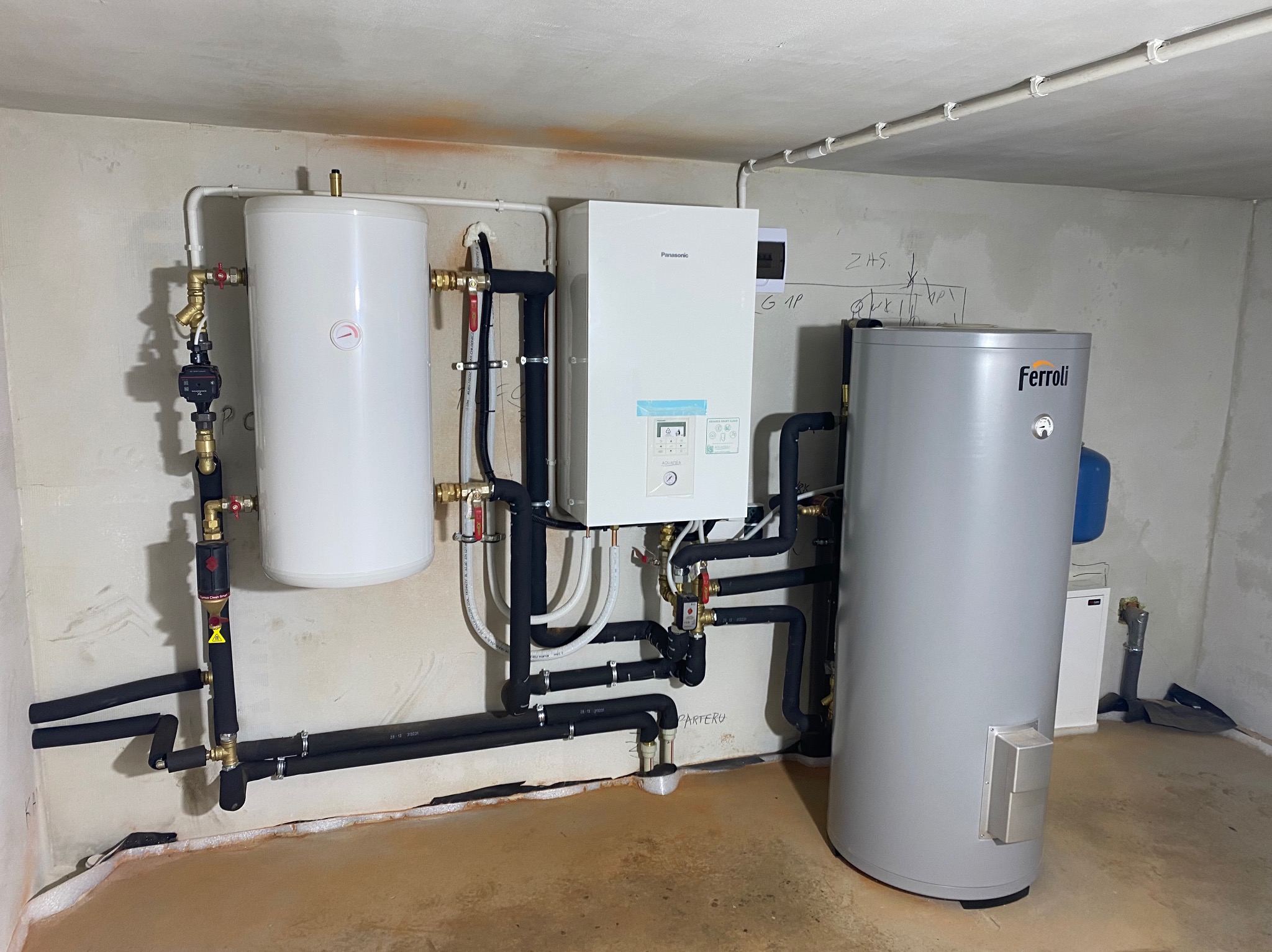 Zestaw urządzeń grzewczych: biały zbiornik, jednostka Panasonic Aquarea i szary zasobnik Ferroli, połączone rurami w piwnicy z surowymi ścianami.