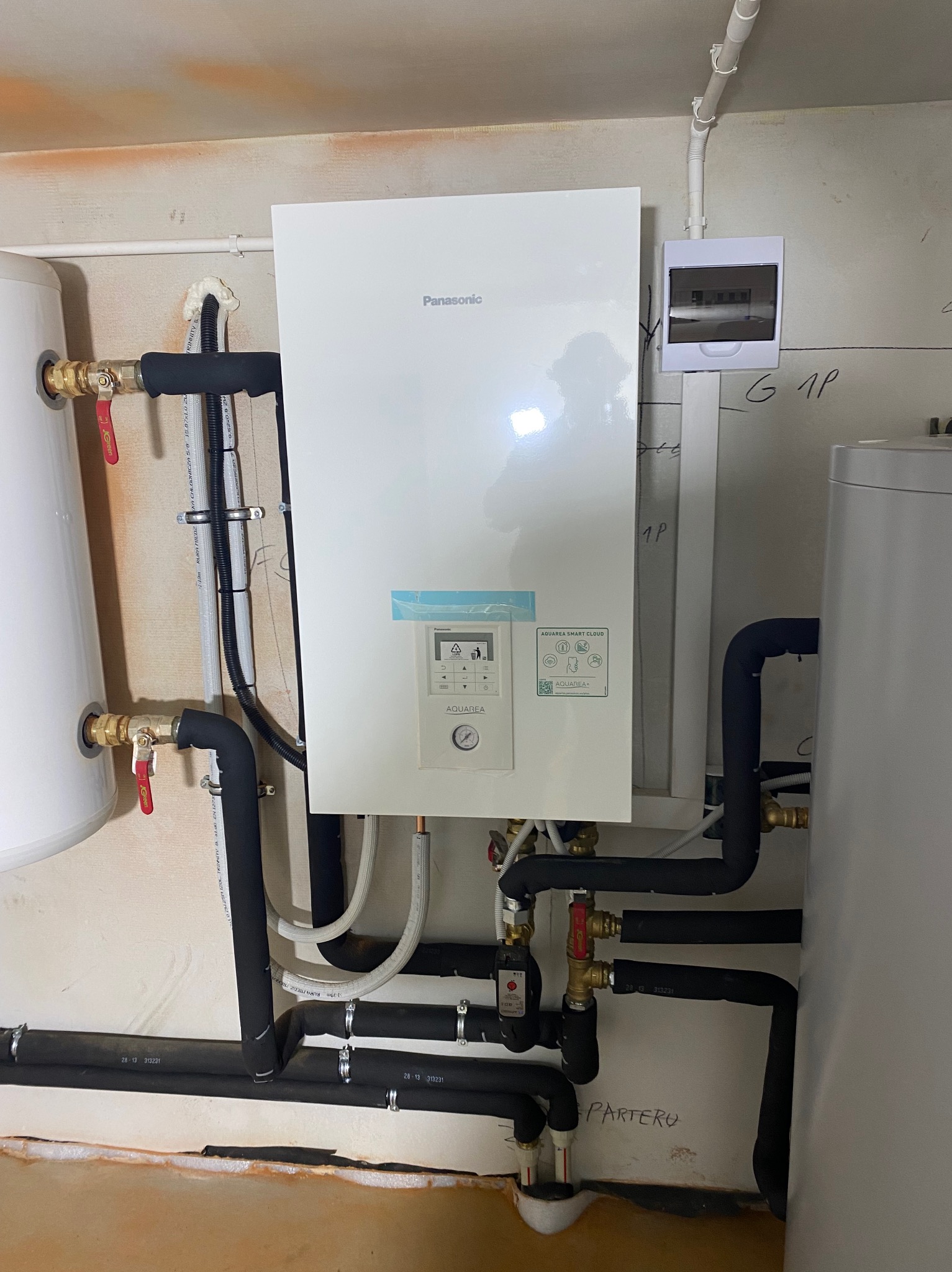 Biała pompa ciepła Panasonic Aquarea zamontowana na ścianie, widoczne rury w izolacji, zawory, zbiornik buforowy i skrzynka elektryczna, instalacja w piwnicy, ściana z napisem 'PARTERU'.