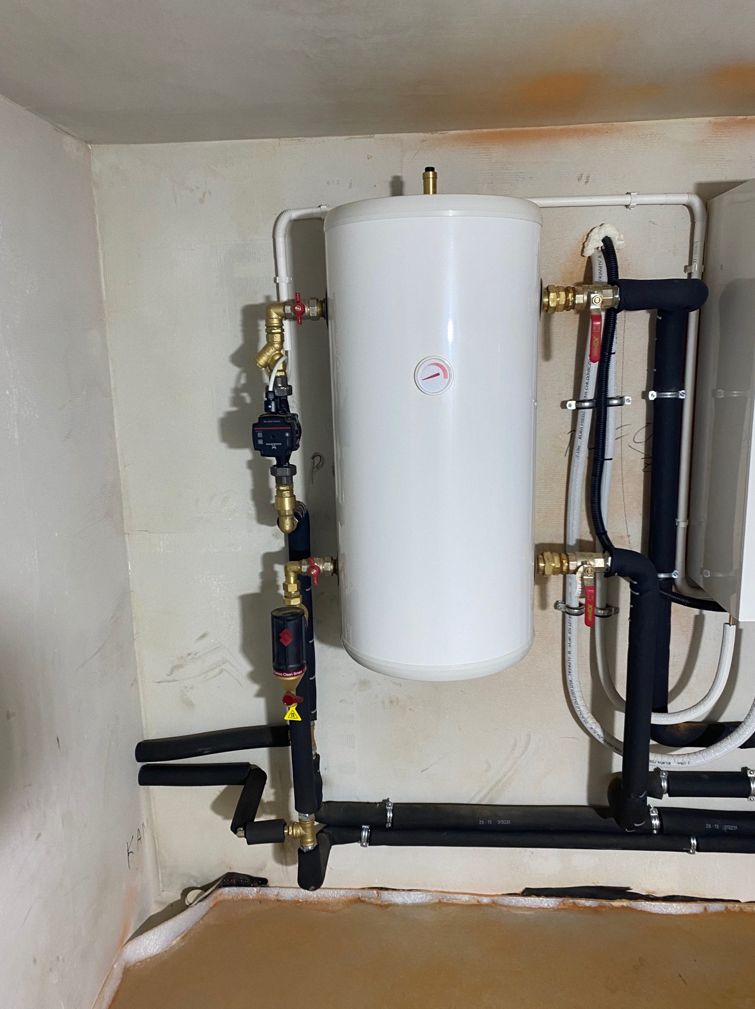 Biały zasobnik ciepłej wody użytkowej z czarnymi rurami izolowanymi termicznie i mosiężnymi zaworami w trakcie montażu w Jedwabnie, widoczne ślady po remoncie na ścianach.
