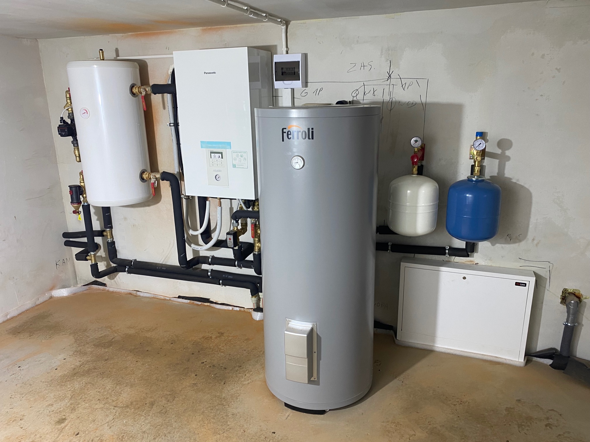 Instalacja grzewcza w piwnicy z kotłem Ferroli, zasobnikiem ciepłej wody Panasonic i rurami doprowadzającymi, widoczne manometry i schemat instalacji na ścianie.