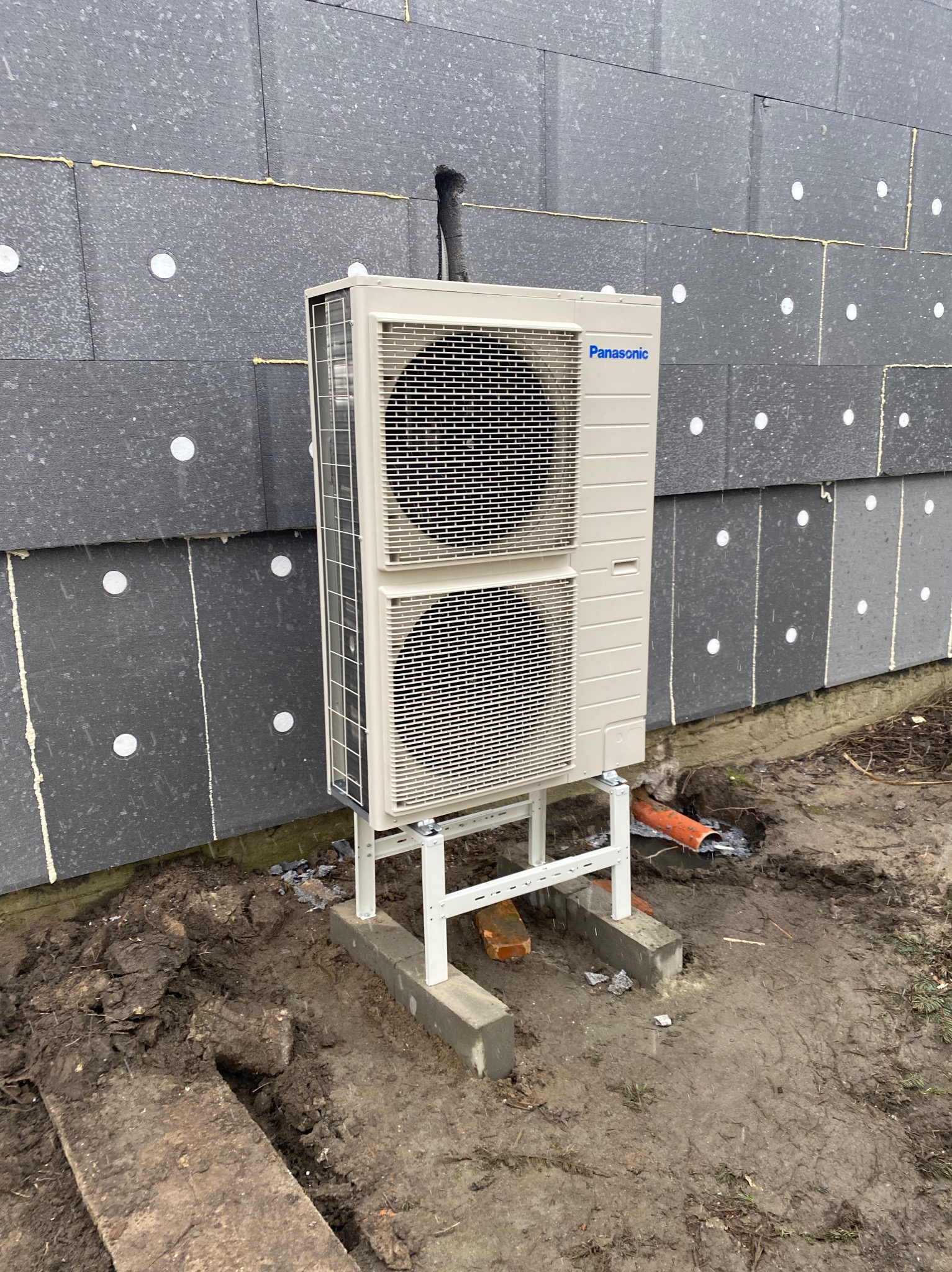 Zewnętrzna jednostka klimatyzacji Panasonic zamontowana na betonowej podstawie przy ocieplonej styropianem ścianie budynku z widoczną rurą odprowadzającą skropliny.