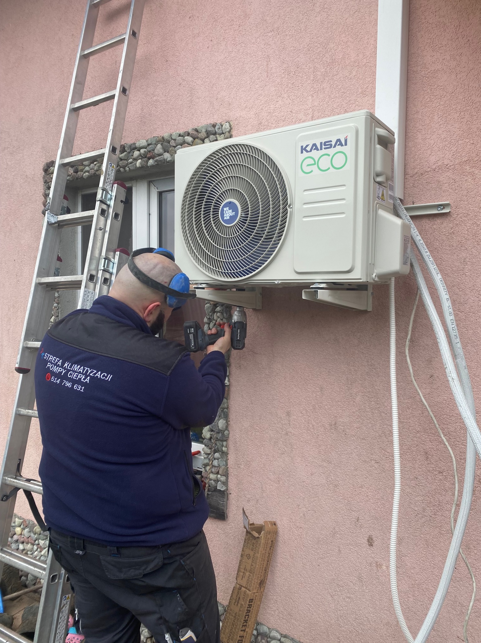Monteraż klimatyzatora Kaisai ECO na elewacji budynku, instalator w niebieskiej bluzie firmowej z numerem telefonu wierci w ścianie, drabina oparta o ścianę, widoczne przewody i karton po uchwycie.
