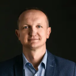 Portret mężczyzny w niebieskiej marynarce i wzorzystej koszuli na ciemnym tle, uśmiechający się, z widocznymi zmarszczkami wokół oczu.