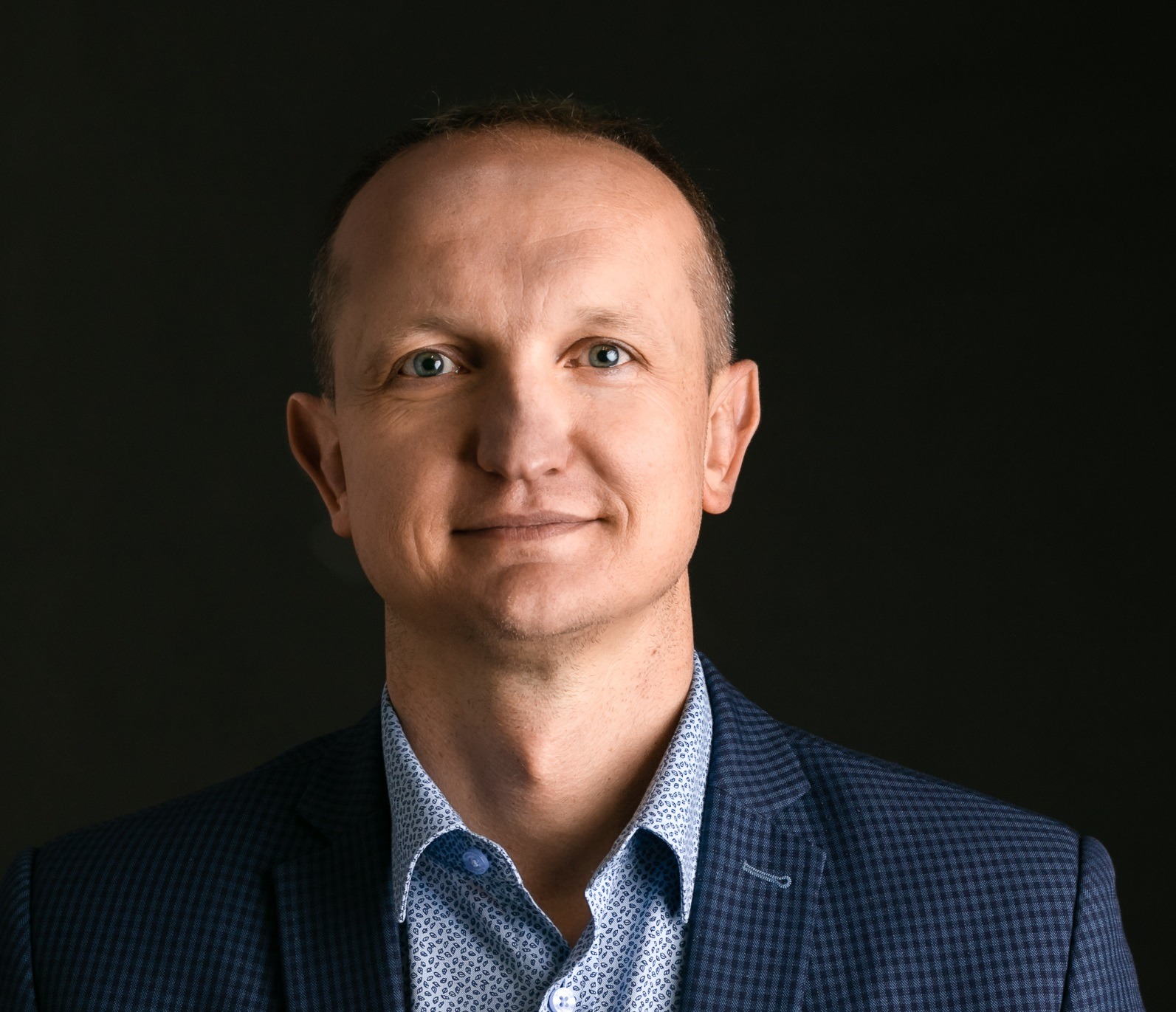 Portret mężczyzny w niebieskiej marynarce i wzorzystej koszuli na ciemnym tle, uśmiechający się, z widocznymi zmarszczkami wokół oczu.