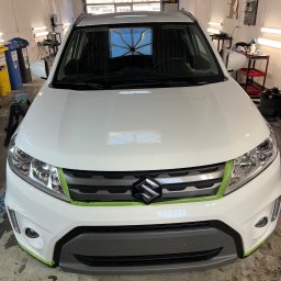 Yauheni Mardasevich - Biały samochód Suzuki zabezpieczony taśmą malarską przed zabiegami w warsztacie, widok z przodu na maskę i grill. W tle widoczne narzędzia i wyposażenie warsztatowe.