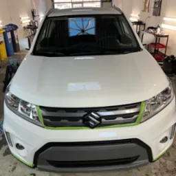 Biały samochód Suzuki zabezpieczony taśmą malarską przed zabiegami w warsztacie, widok z przodu na maskę i grill. W tle widoczne narzędzia i wyposażenie warsztatowe.