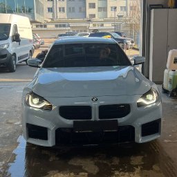 Yauheni Mardasevich - Białe BMW M2 stoi wewnątrz myjni, widoczne mokre ślady po umyciu na posadzce, w tle budynek biurowy i inne samochody.