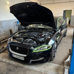 Yauheni Mardasevich - Czarny Jaguar XJ z otwartą maską, zabezpieczony taśmą malarską wokół reflektorów, silnik widoczny, na podnośniku w warsztacie.