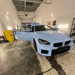 Yauheni Mardasevich - Jasnoniebieski samochód z otwartymi drzwiami w warsztacie, widoczny żółty kosz na śmieci i narzędzia na półce w tle.