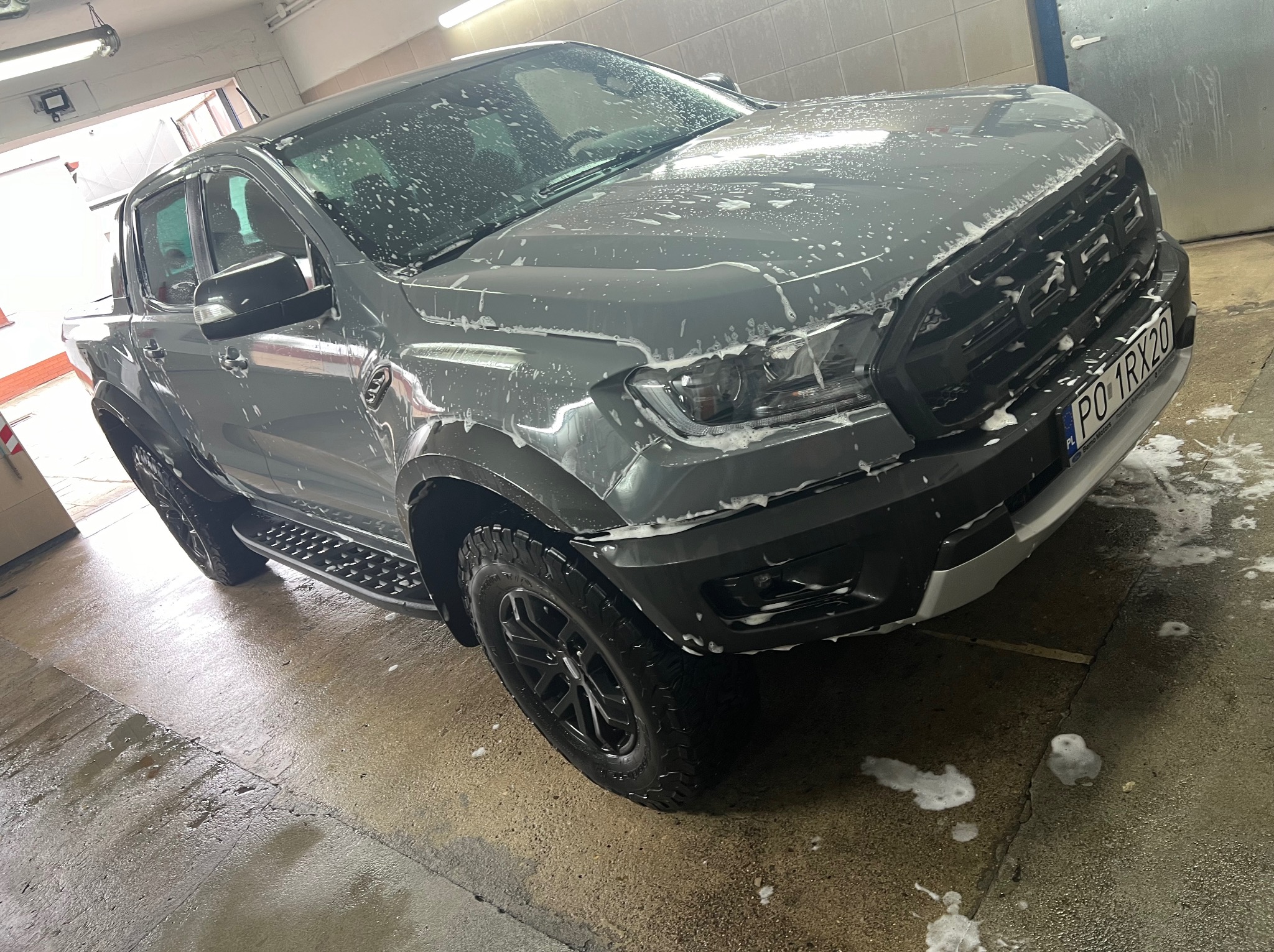 Szary Ford Ranger Raptor pokryty pianą myjącą wewnątrz myjni, widoczne tablice rejestracyjne z polskim numerem, mokra posadzka.