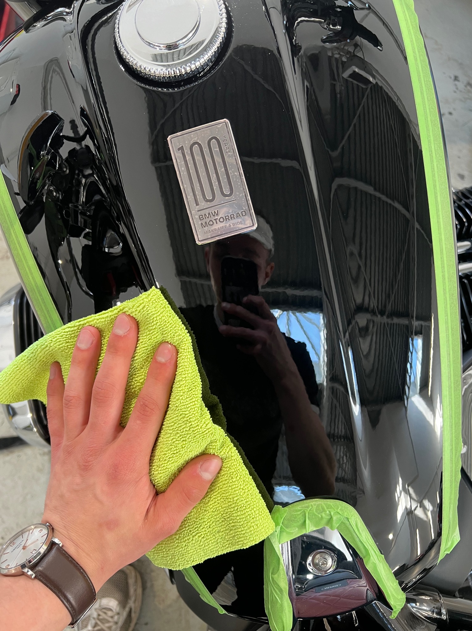 Czyszczenie lakieru motocykla BMW, zabezpieczonego taśmą, miękką, zieloną ściereczką z mikrofibry, widoczny zegarek na ręce osoby wykonującej.