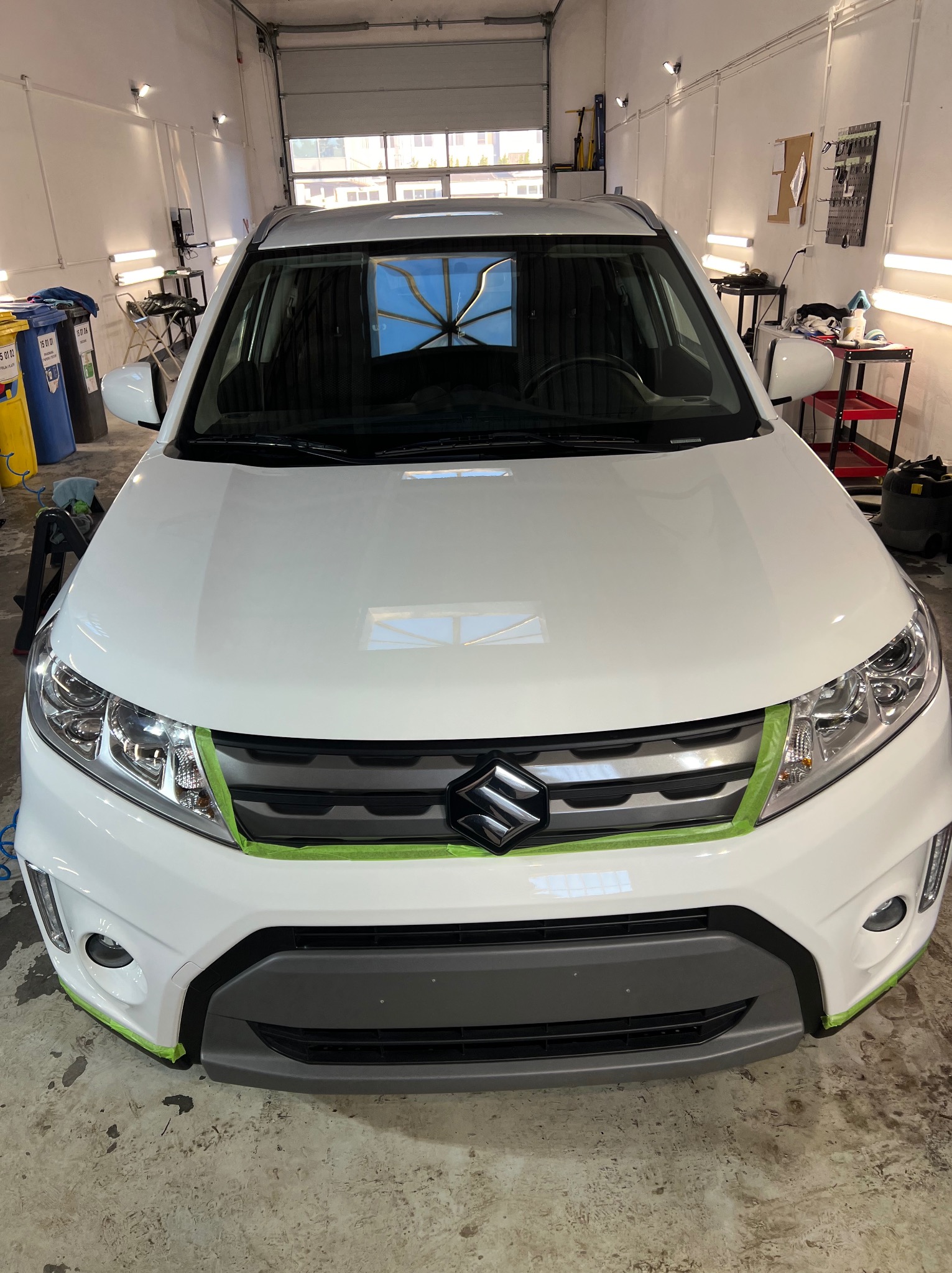 Biały samochód Suzuki zabezpieczony taśmą malarską przed zabiegami w warsztacie, widok z przodu na maskę i grill. W tle widoczne narzędzia i wyposażenie warsztatowe.