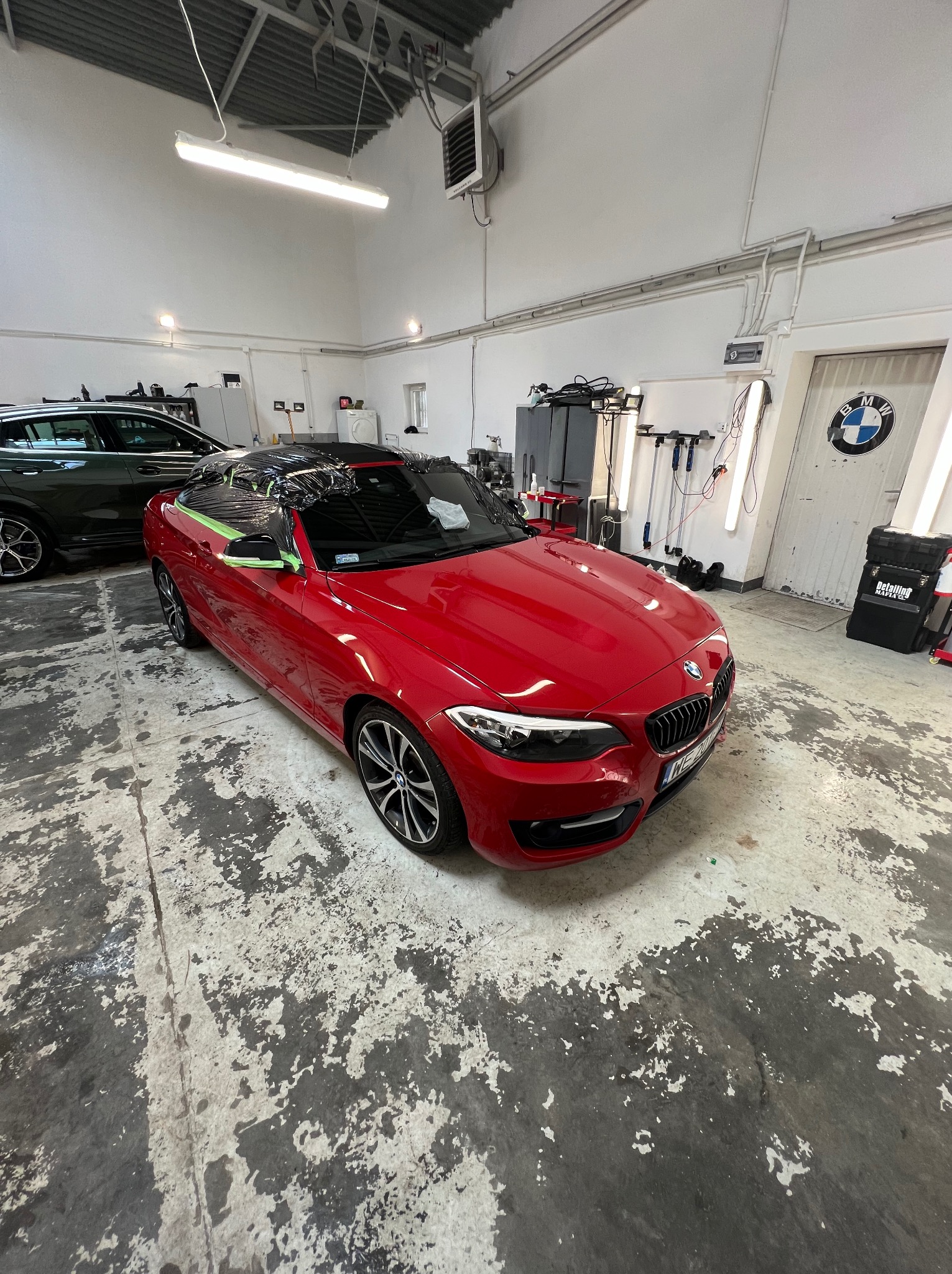 Czerwone BMW serii 2 Cabrio zabezpieczone folią ochronną i taśmą malarską przed oklejaniem, widok z przodu w warsztacie detailingowym z betonową podłogą i logiem BMW na drzwiach w tle.